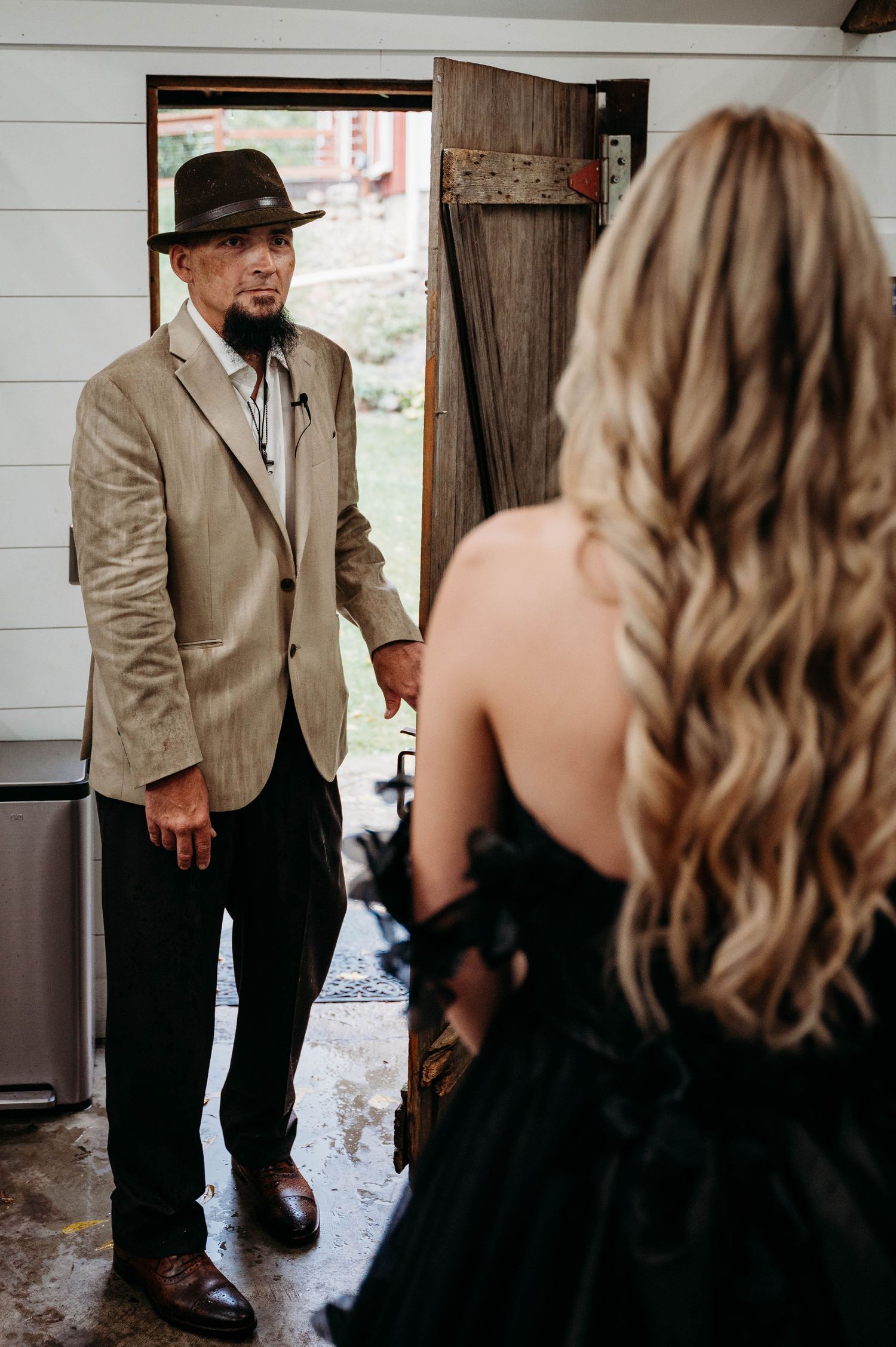 Emotional moment at a rustic doorway between a gentleman in a beige suit and hat and someone in a black formal dress.