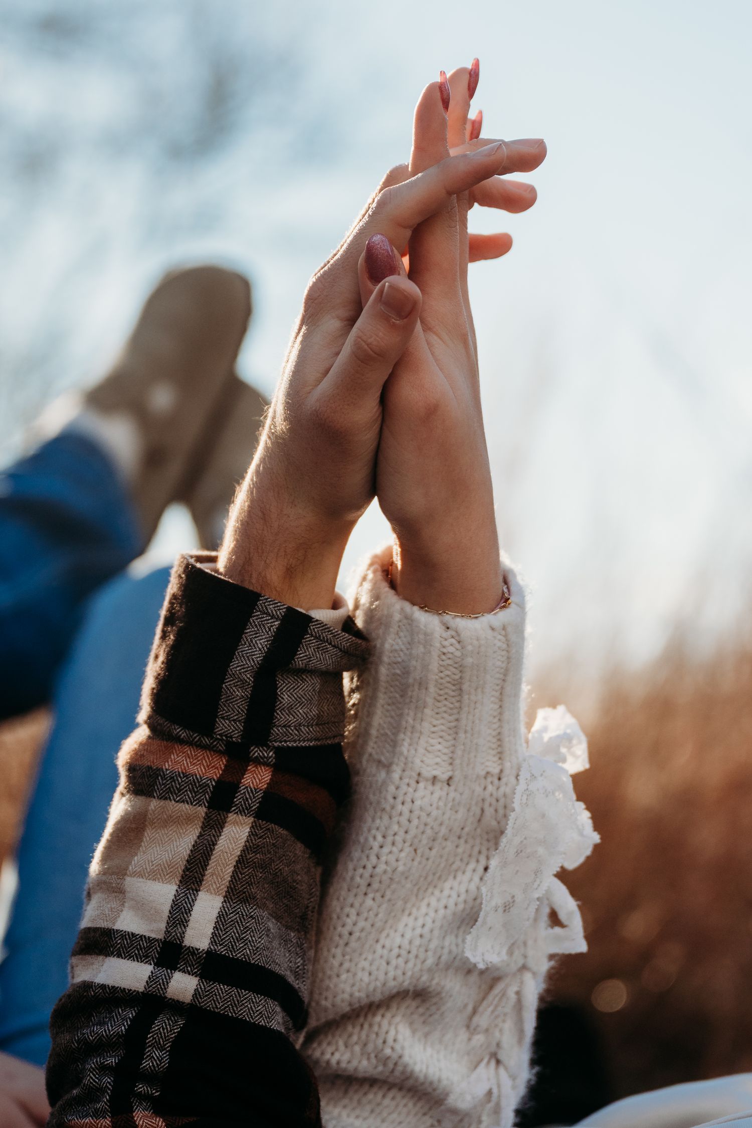 Two hands reach upward wearing contrasting sleeves - one in plaid pattern and one in white knit - against a blurry sky.