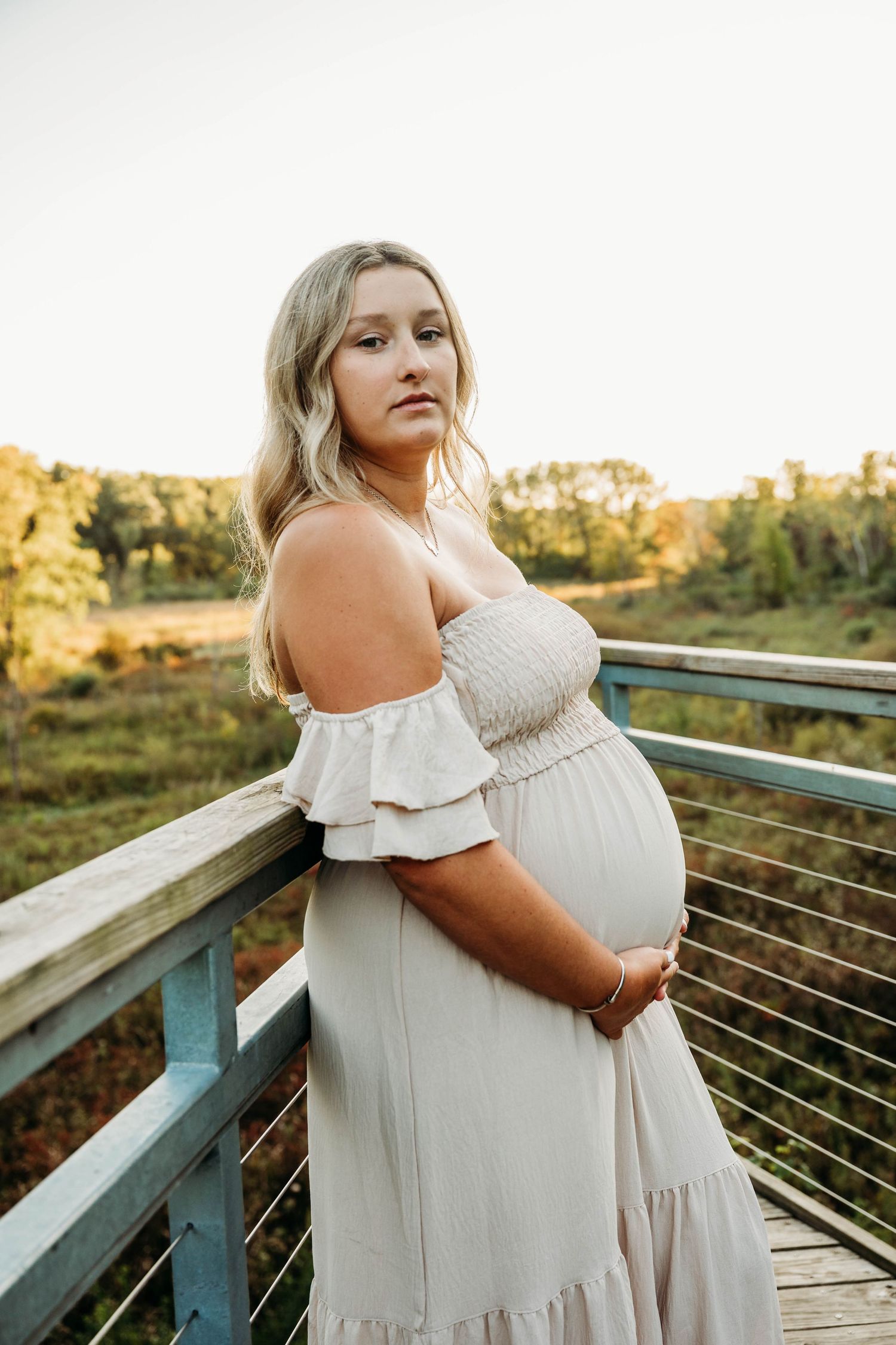 Expectant mother in flowing white dress poses on wooden deck during golden hour sunset.