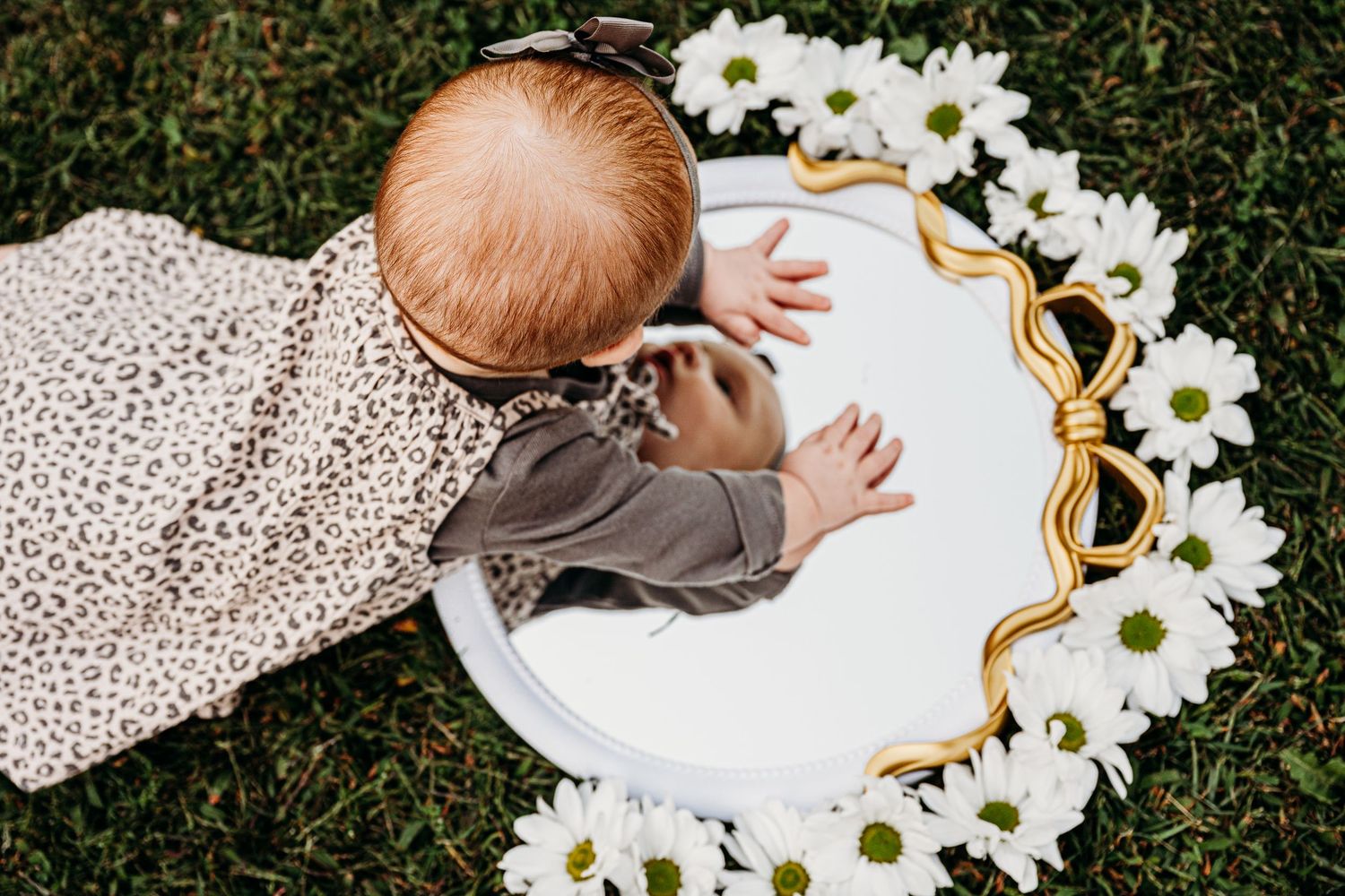 Baby looking down at round mirror surrounded by white flowers on grass.