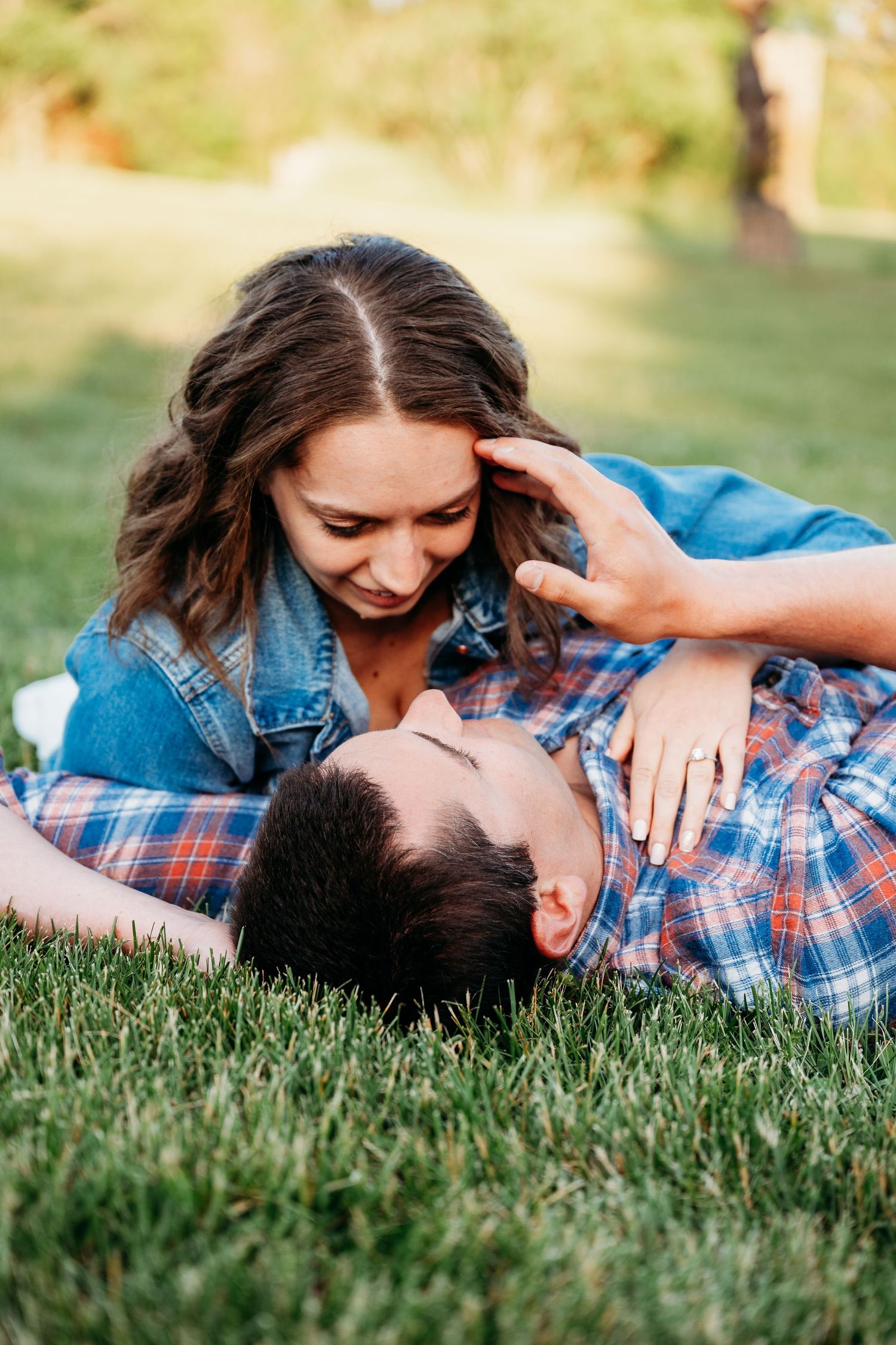 Couple relaxing on green grass, woman smiling down at man lying in her lap, both wearing casual denim and plaid shirts.