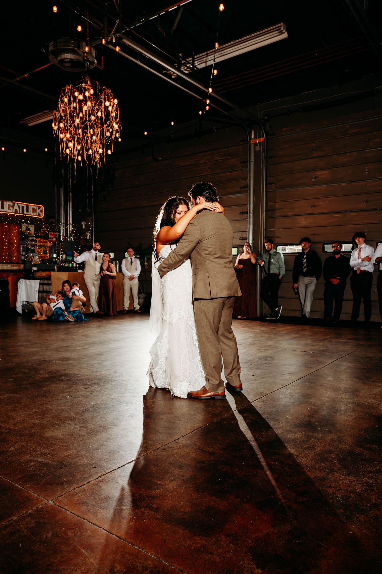 Bride and groom dancing alone on a wooden dance floor under warm string lights and chandelier in a rustic barn venue.