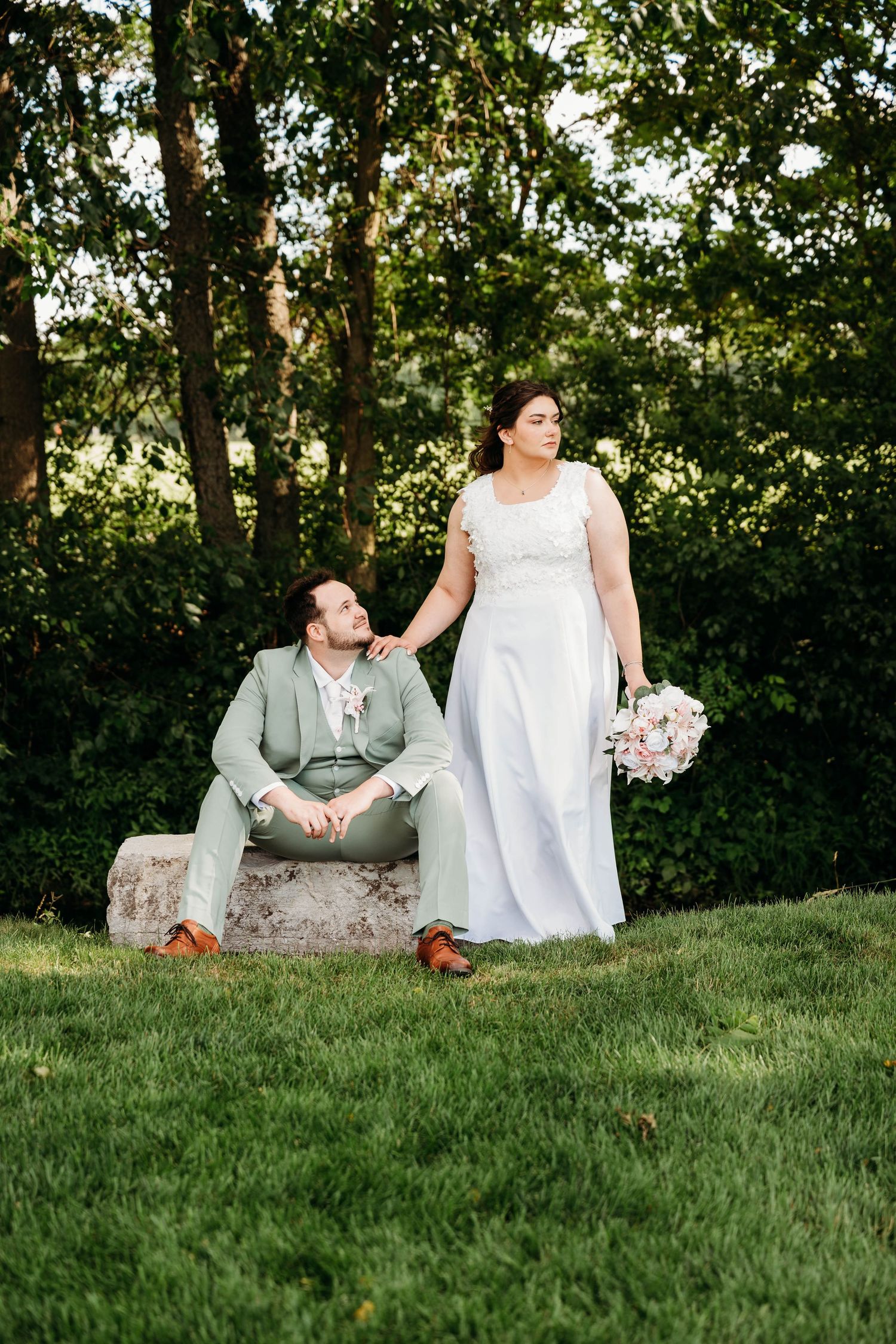 Bride in white gown holds bouquet while groom in mint suit sits on stone bench outdoors.