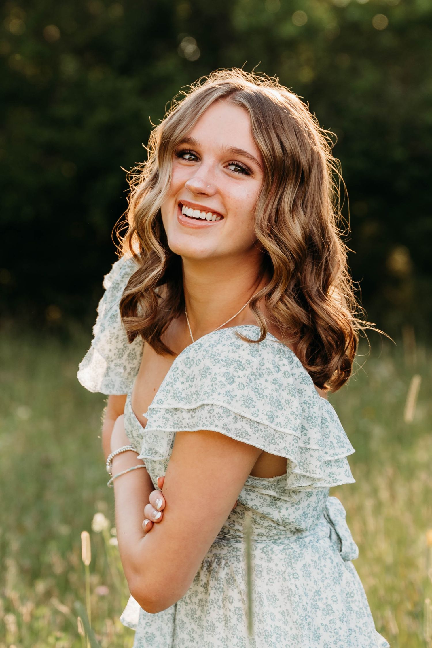Smiling young woman in a floral ruffle dress standing in a sunlit field with lush green trees in the background.