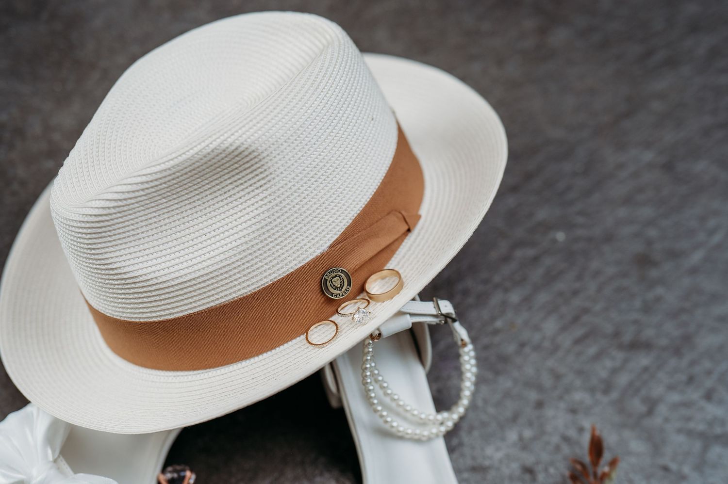 White Panama hat with brown band and gold accessories, paired with pearl bracelet, on gray background.