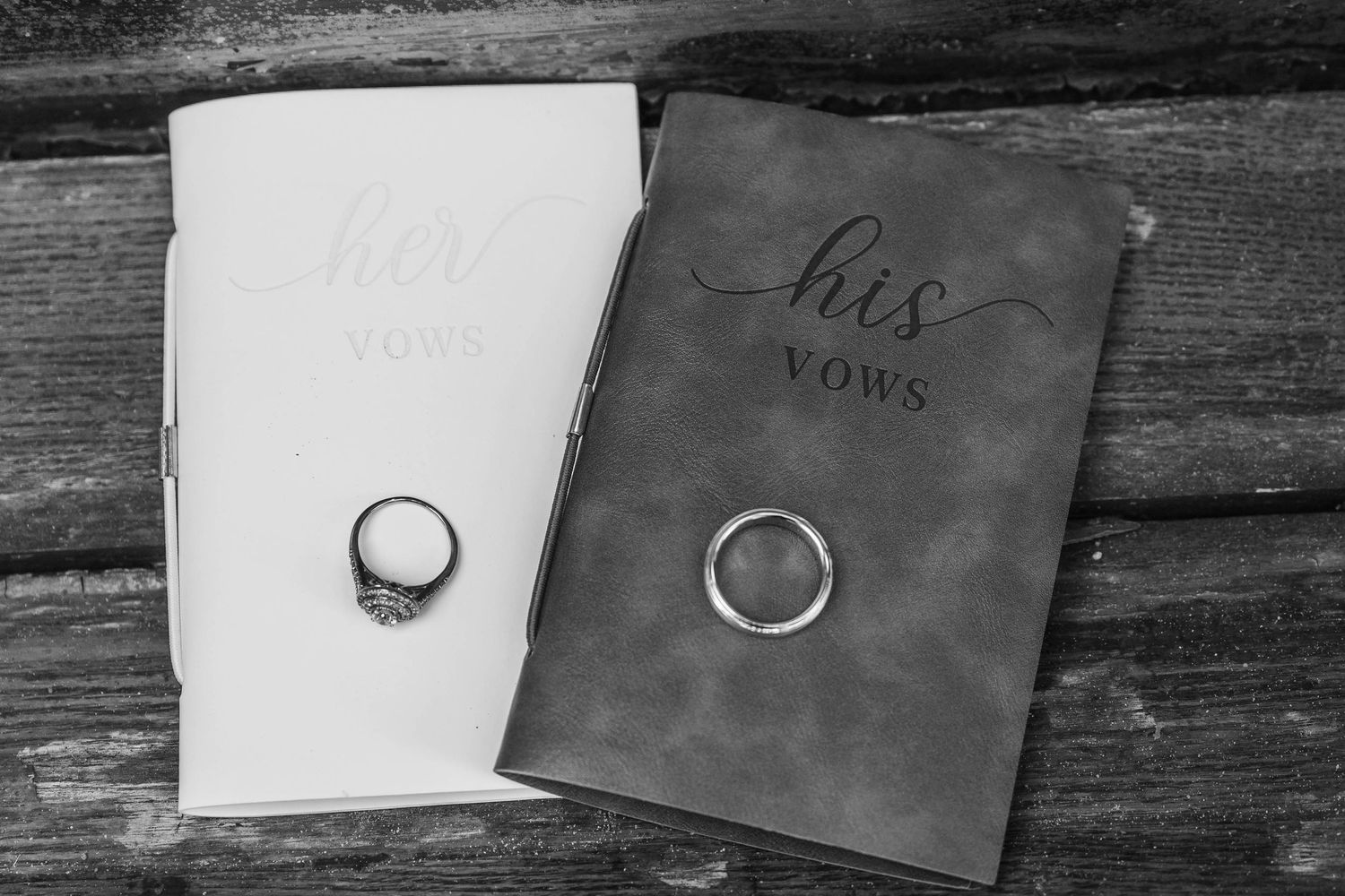 Black and white photo of his and her wedding vow books with matching rings resting on rustic wood.