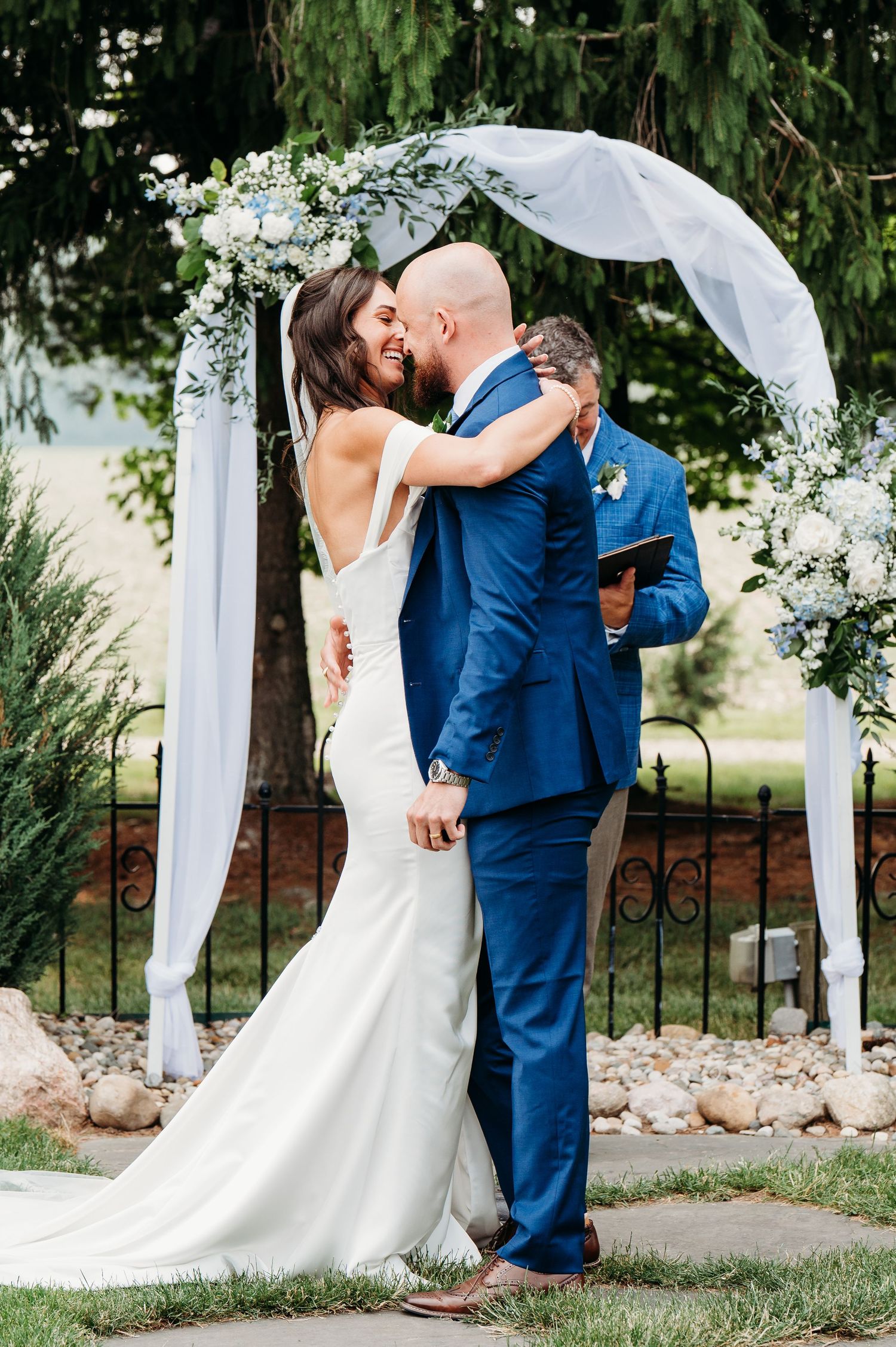 Newlyweds share romantic kiss under floral wedding arch with white fabric and greenery in outdoor garden setting.