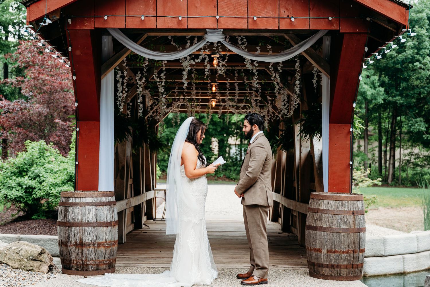Bride and groom exchange vows under a rustic covered bridge decorated with lights and greenery at an outdoor wedding venue.