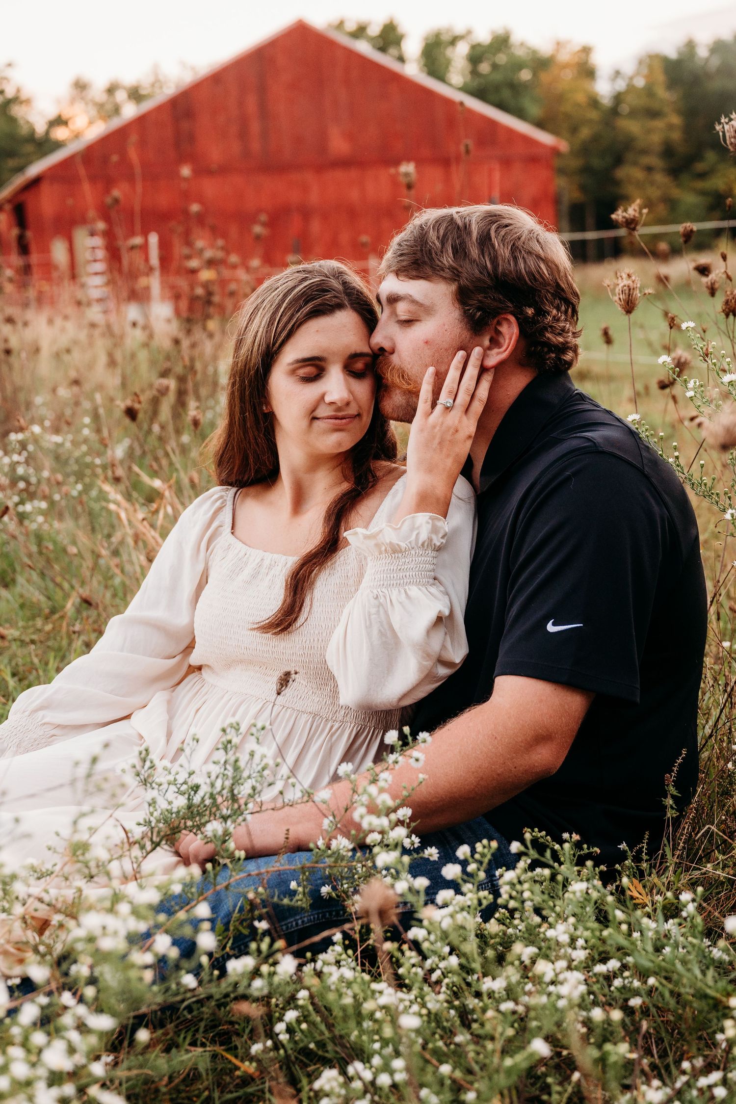 Romantic couple sitting in a wildflower meadow near a red barn during golden hour.