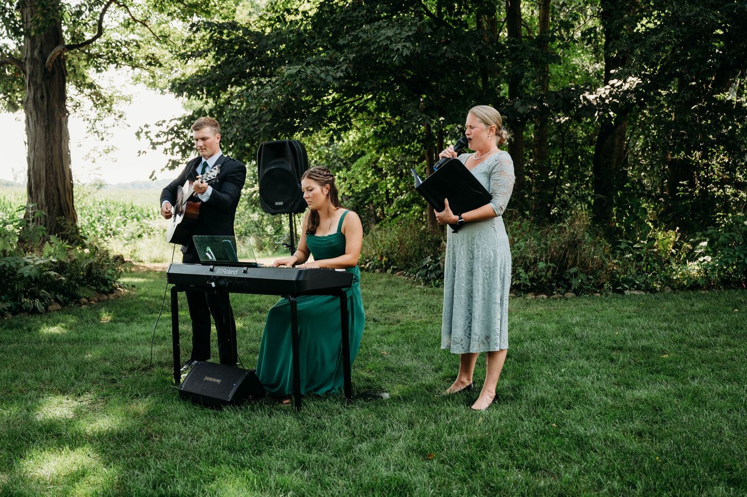 Outdoor wedding musicians perform on green lawn, featuring guitarist, keyboardist, and vocalist in elegant attire.