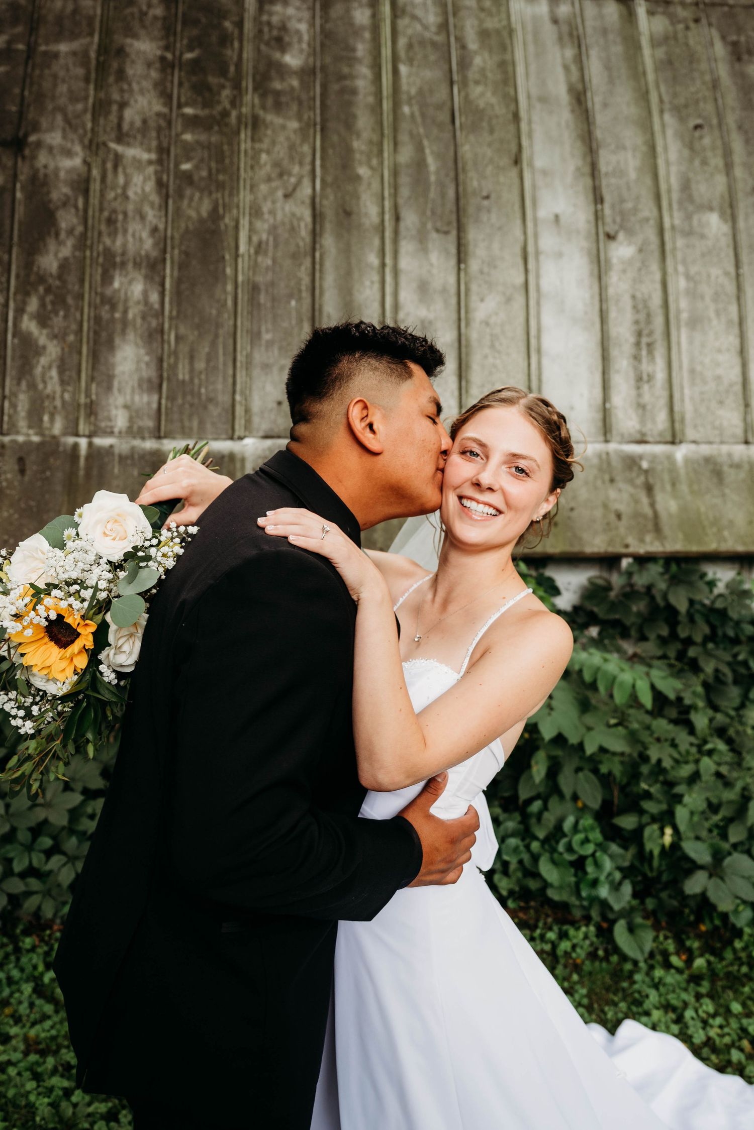 Groom in black suit kisses bride on cheek as she smiles holding sunflower bouquet near rustic barn.