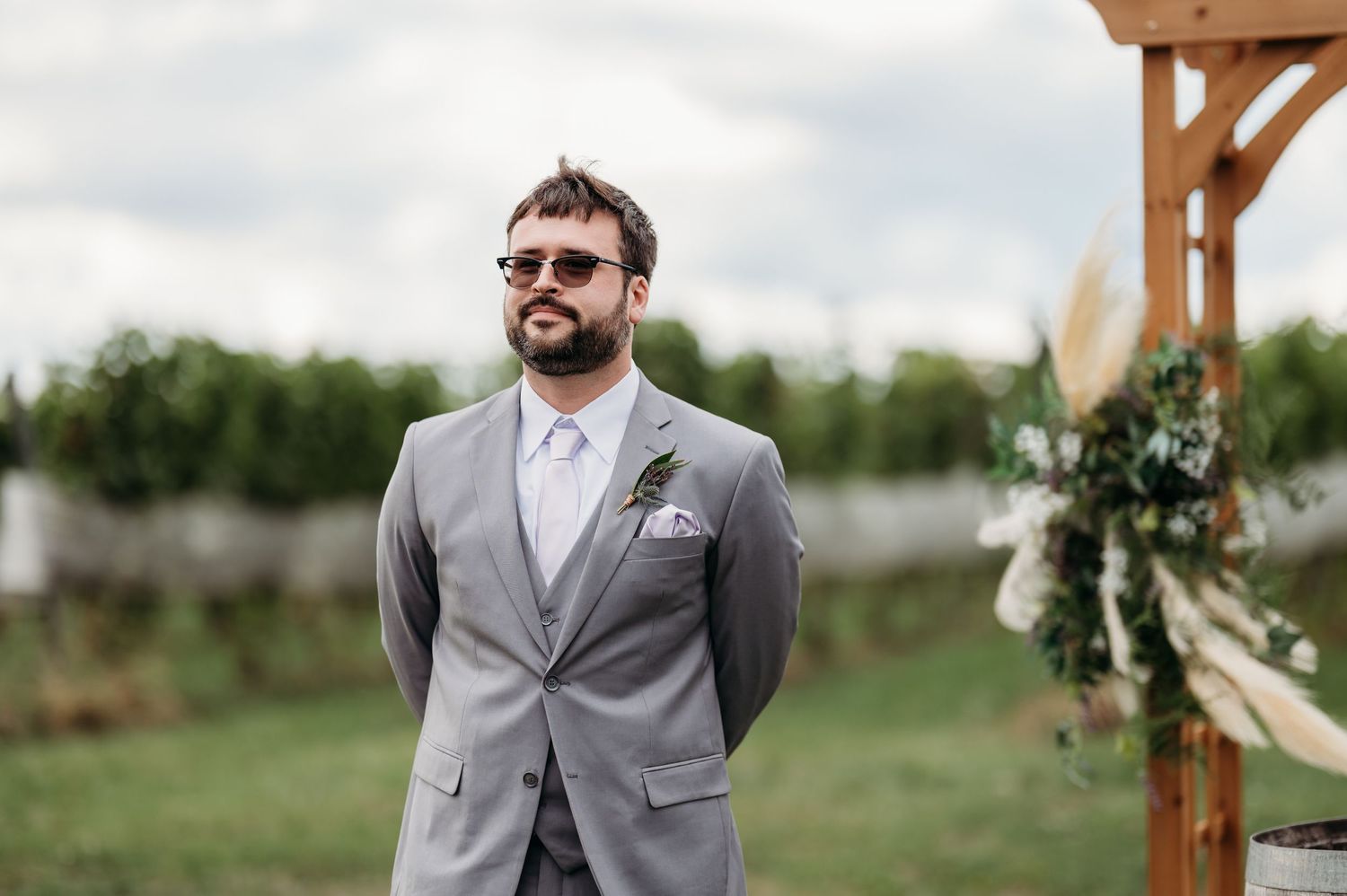 A groom in a light gray suit and sunglasses stands in an outdoor wedding venue with rustic wooden beams.
