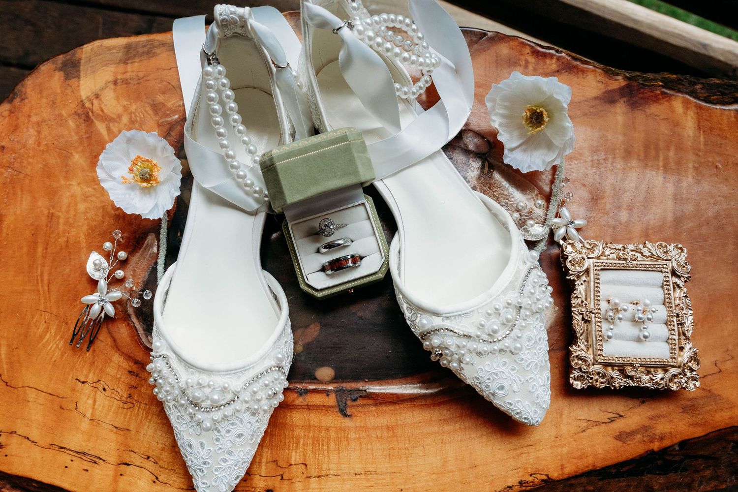 Elegant bridal flat shoes with pearl embellishments displayed with wedding rings, earrings, and white flowers on wood.