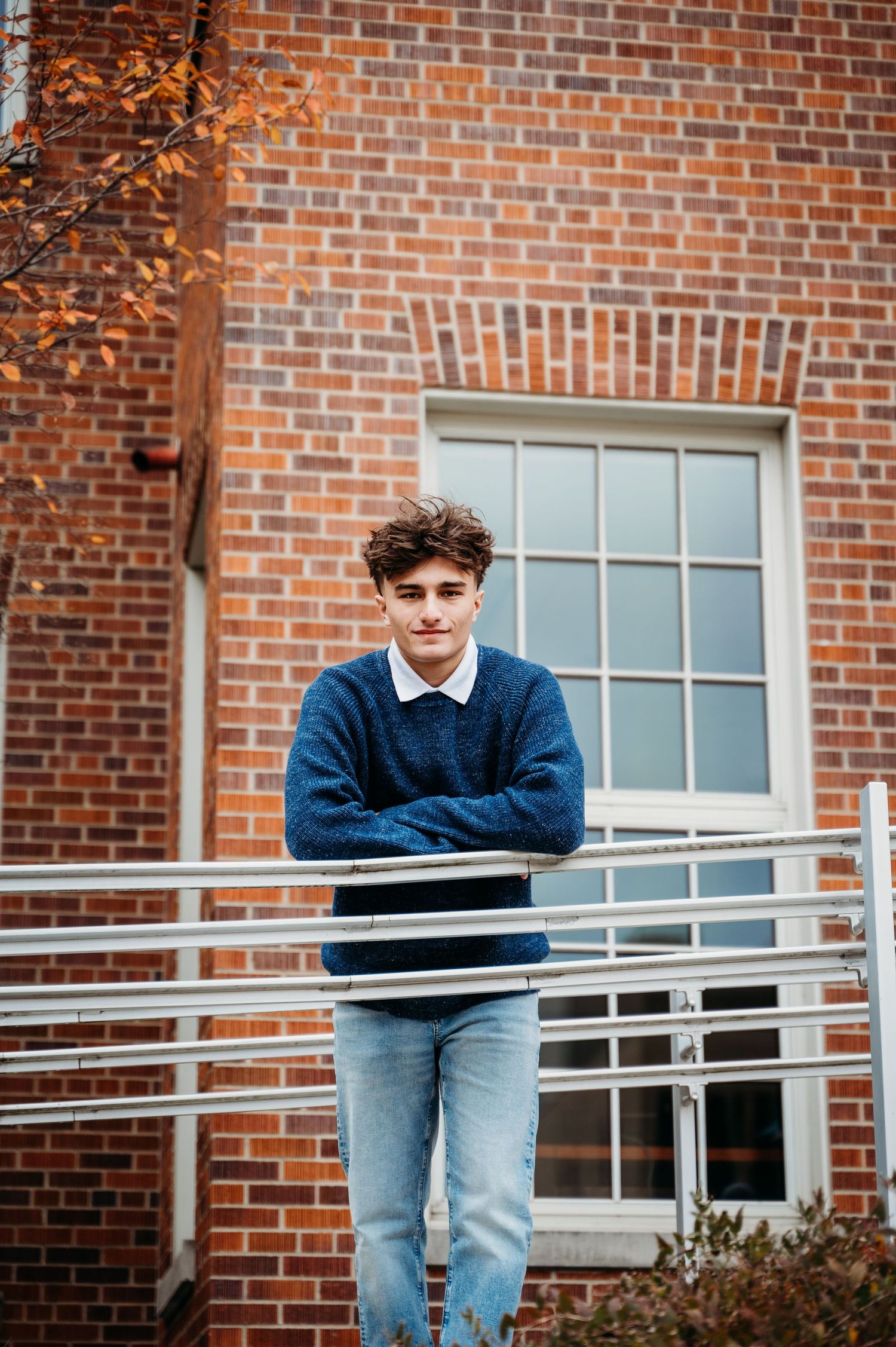 Man in blue sweater leaning against metal railing outside brick building with white window.