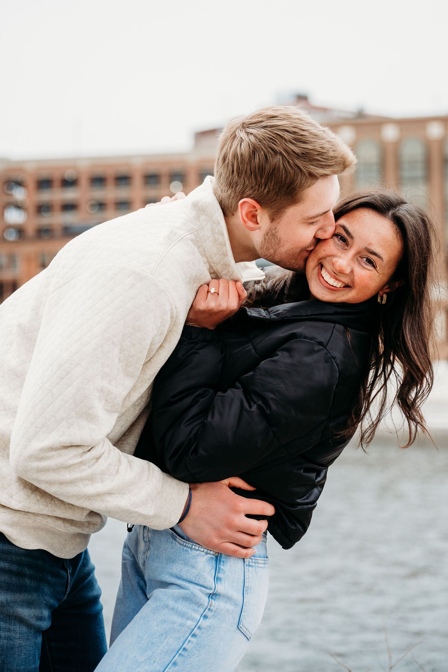 A romantic couple embraces and laughs together outdoors near a brick building on a winter day.