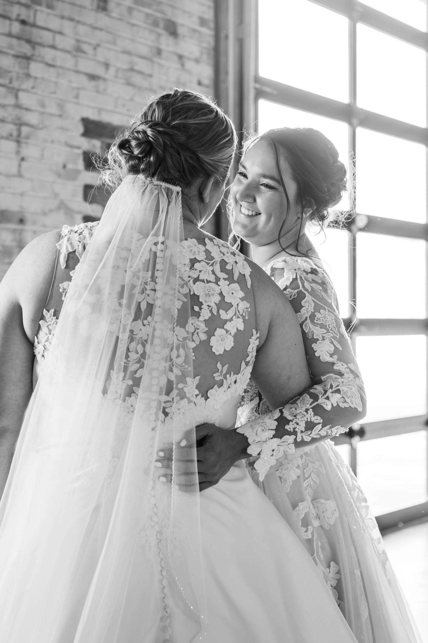 Elegant wedding gowns with lace details and veils captured in black and white against brick wall.