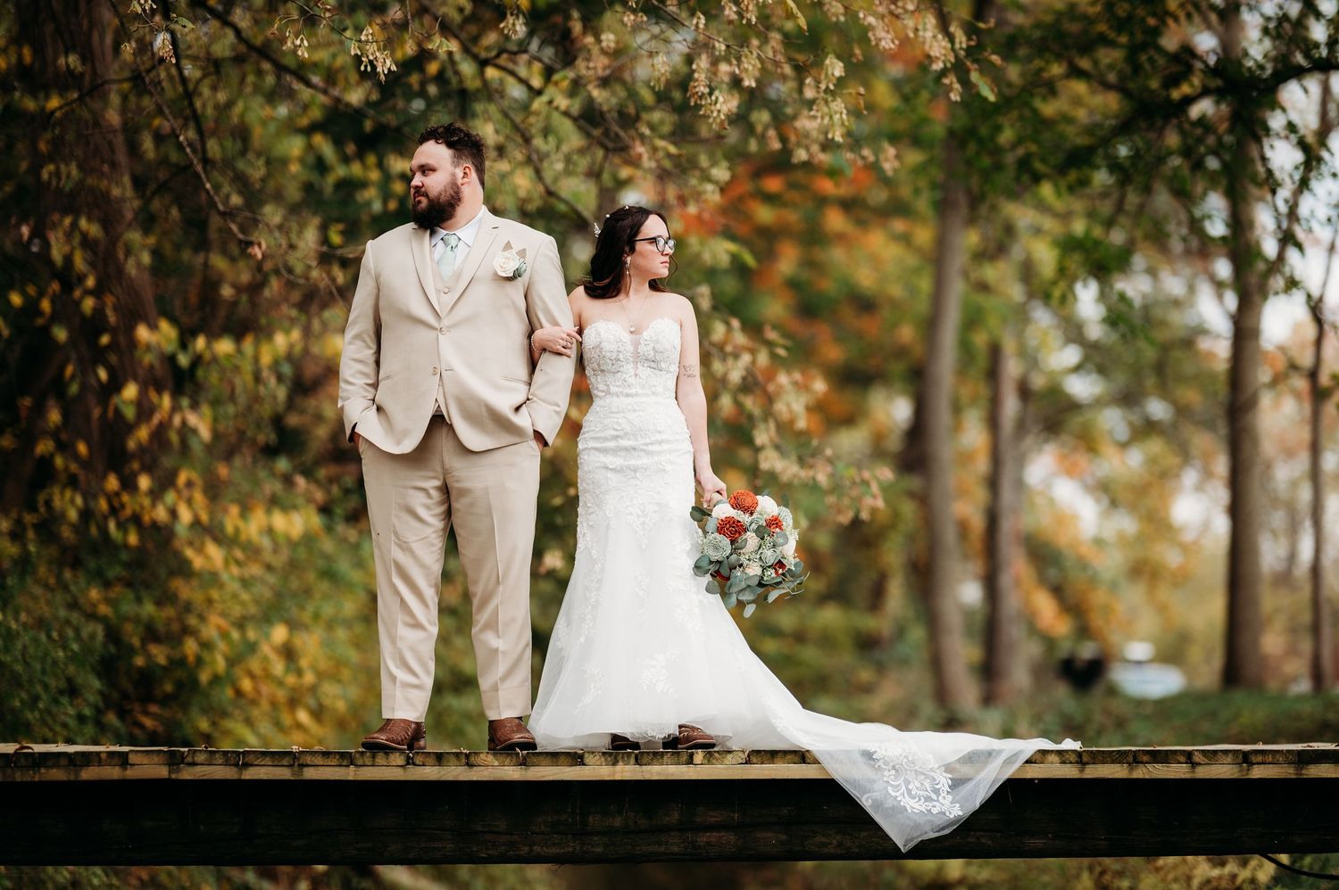 A couple in wedding attire walks through a scenic autumn forest setting with golden leaves and natural light.