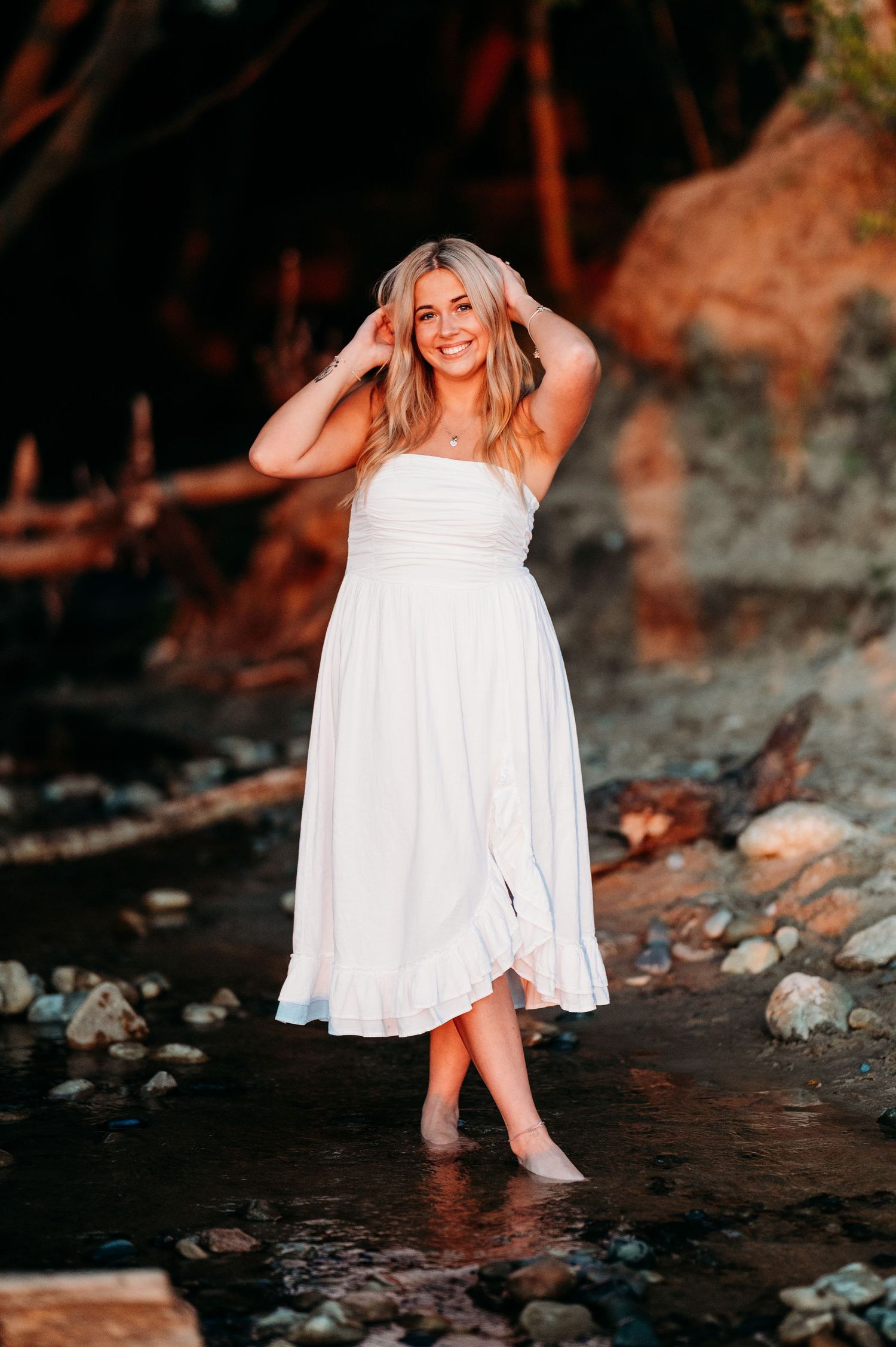 A person in a flowing white sundress stands barefoot in shallow water near rocky cliffs at sunset.