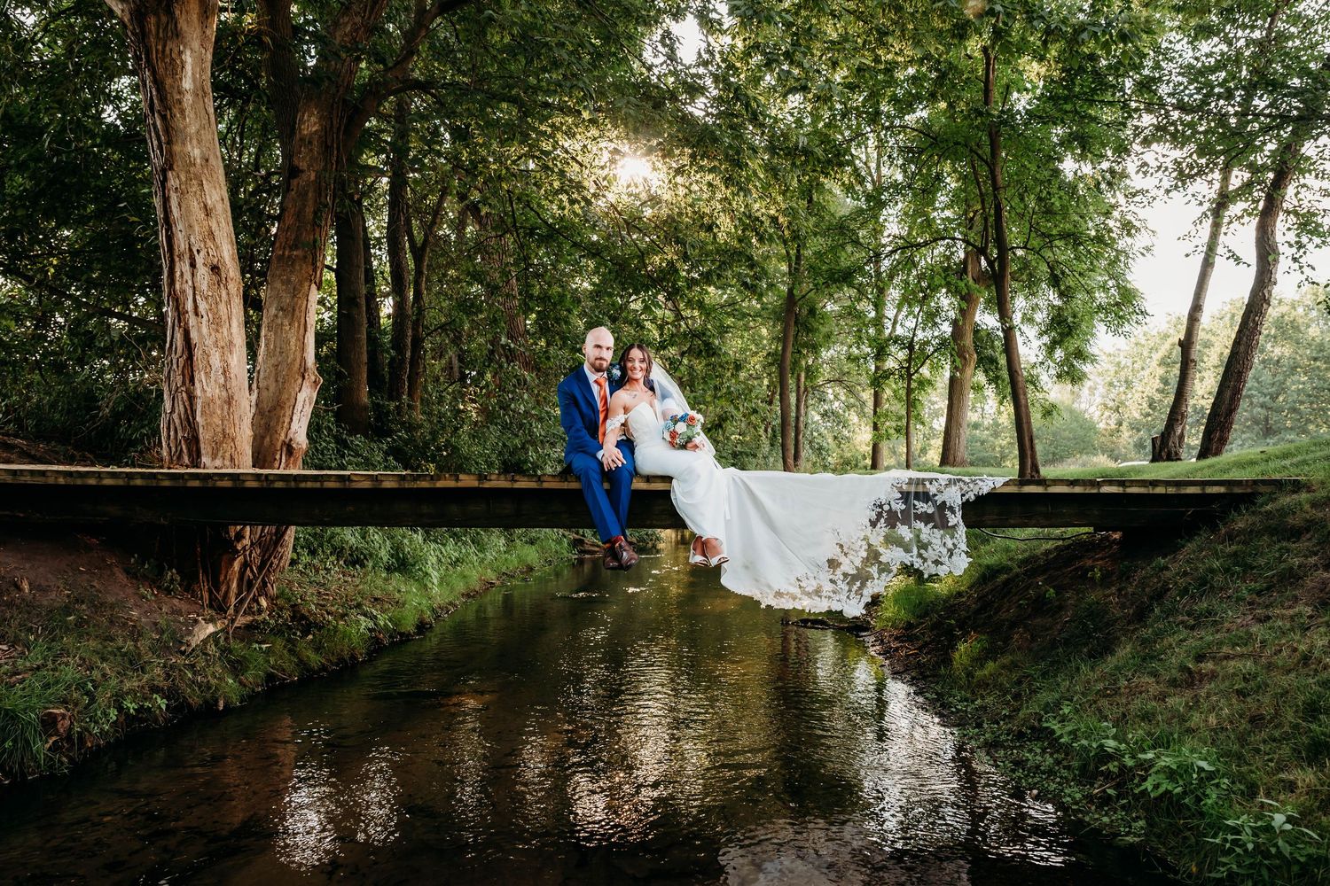 A couple in wedding attire sits together on a wooden bridge surrounded by lush green trees near a tranquil pond.