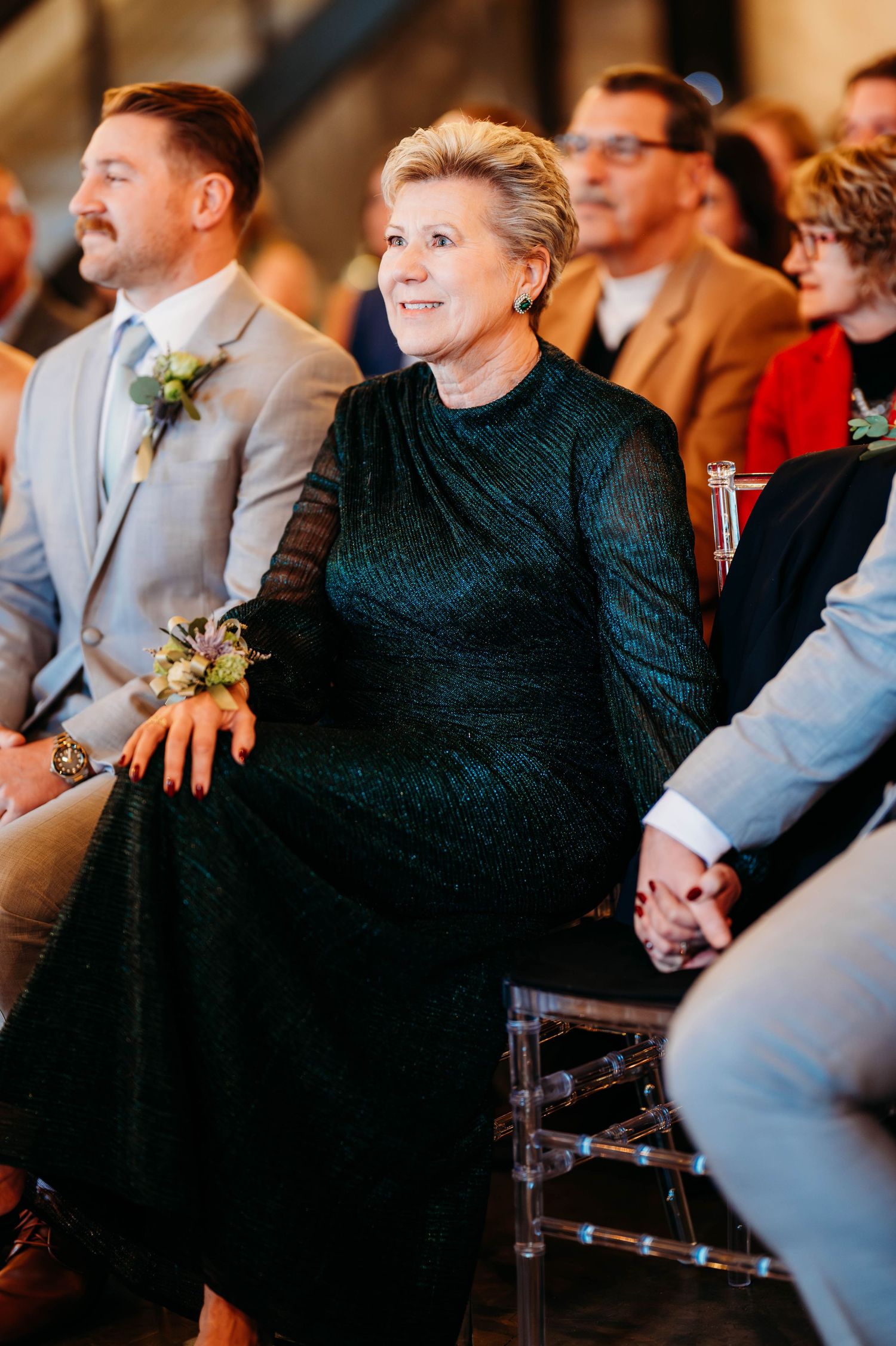 Wedding guests sit in rows during a ceremony, wearing formal attire in dark and neutral colors.