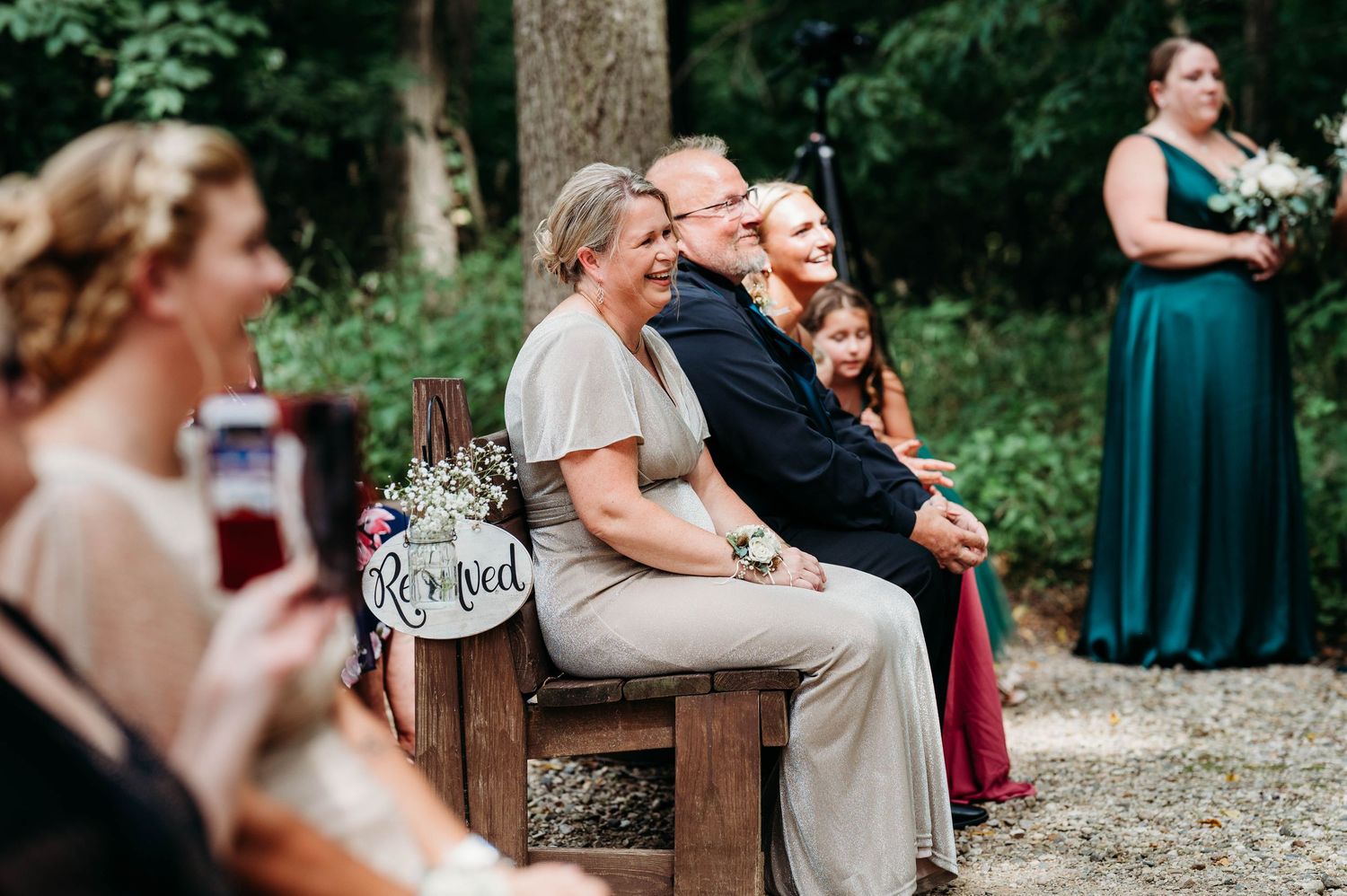 A rustic outdoor wedding ceremony with guests seated on wooden benches surrounded by lush greenery.