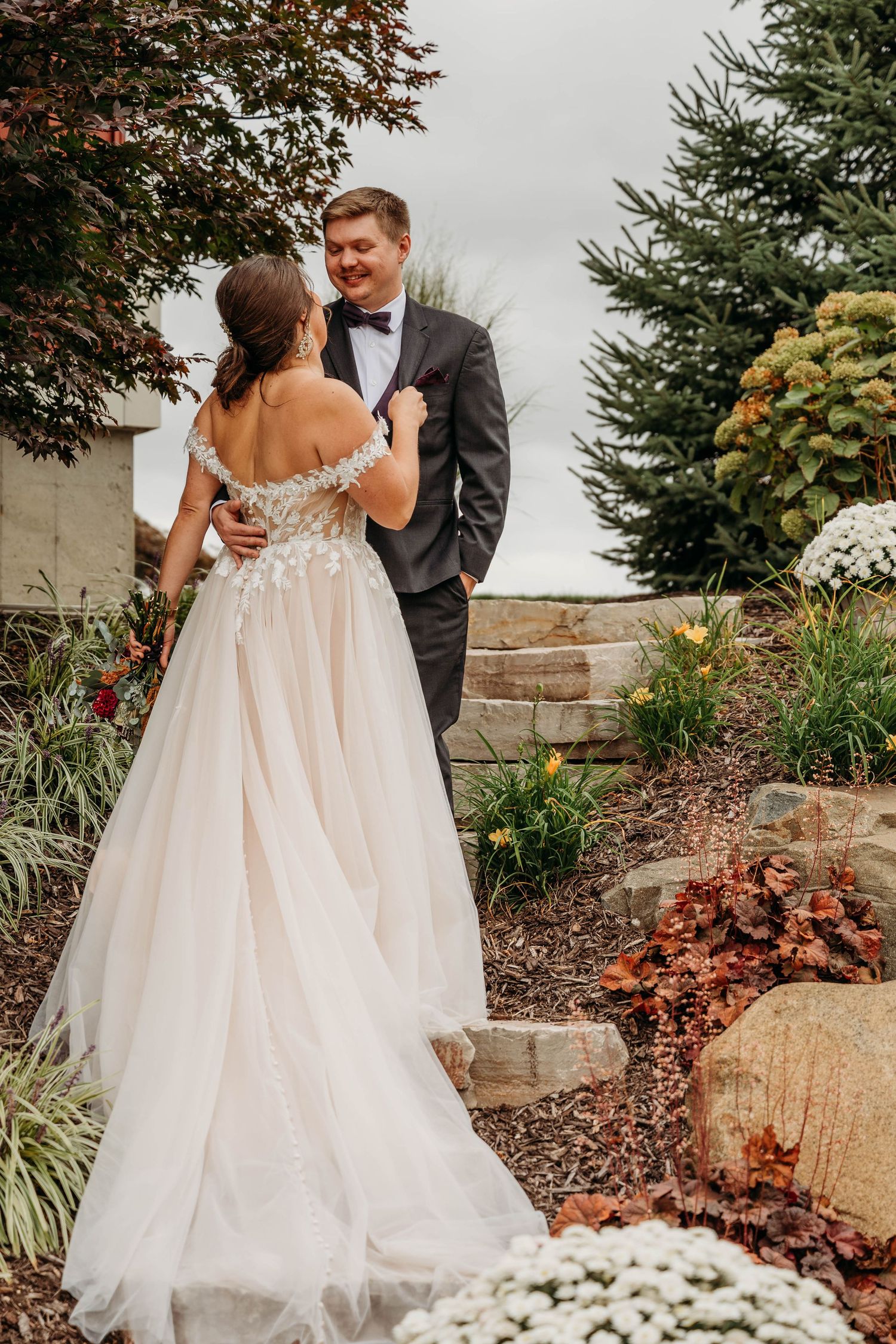 A bride in a flowing tulle gown embraces her partner in a garden setting with stone walls and colorful foliage.