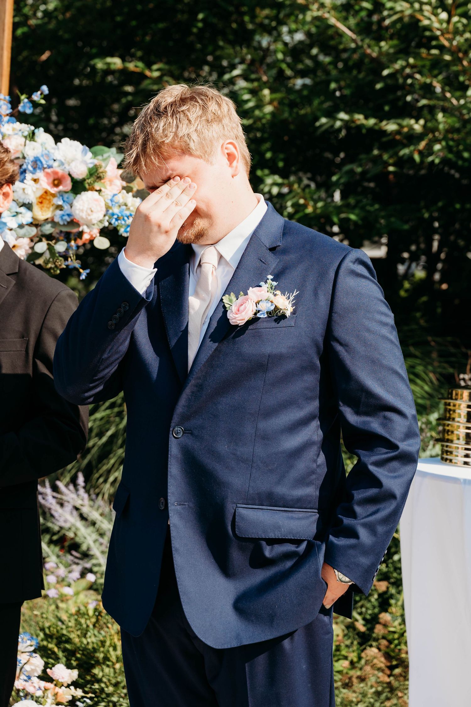 Emotional groom in navy suit with boutonniere wiping tears during outdoor wedding ceremony surrounded by floral arrangements.