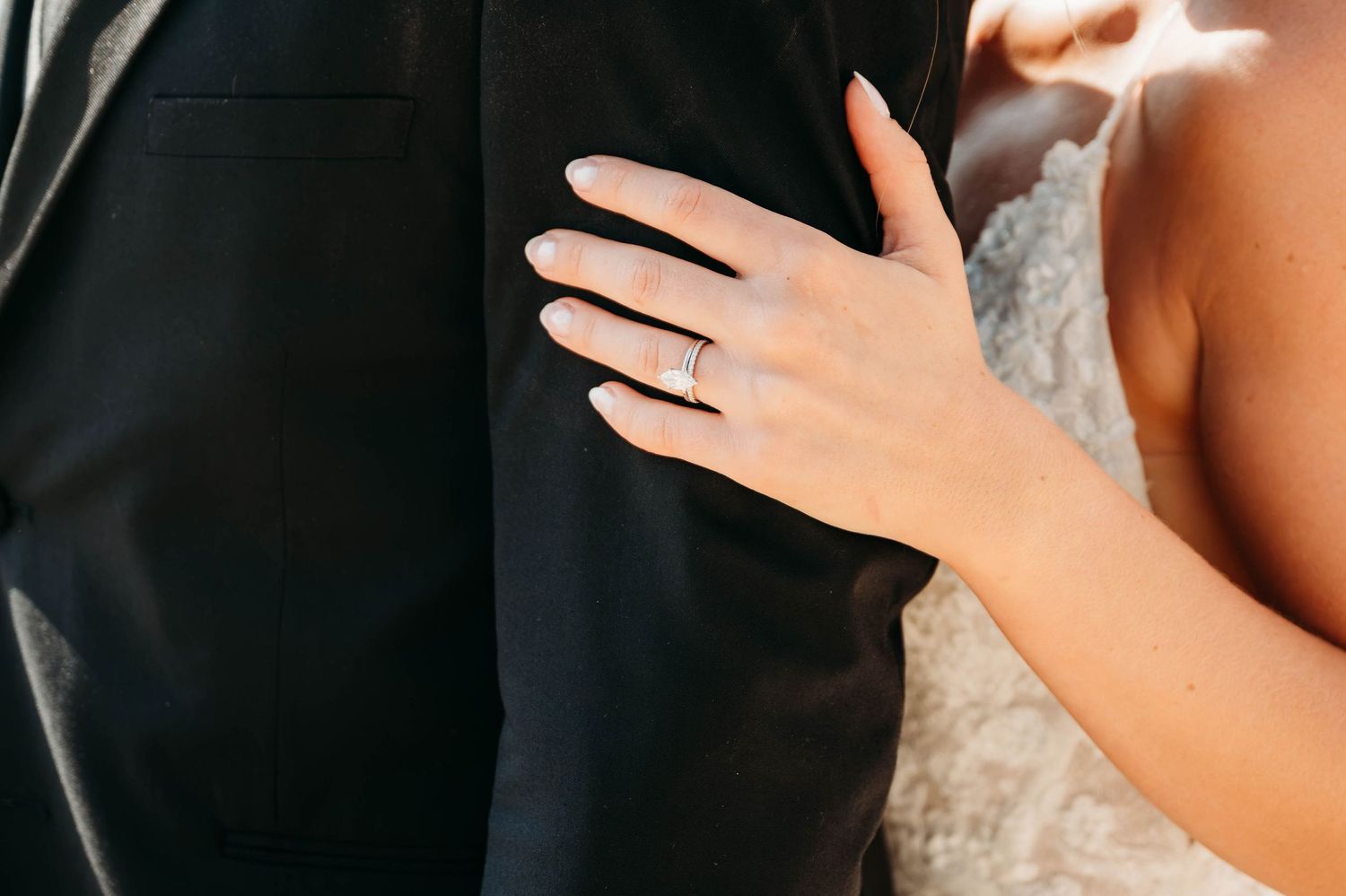 Close-up of a bride's hand with an engagement ring resting on a groom's black suit jacket.