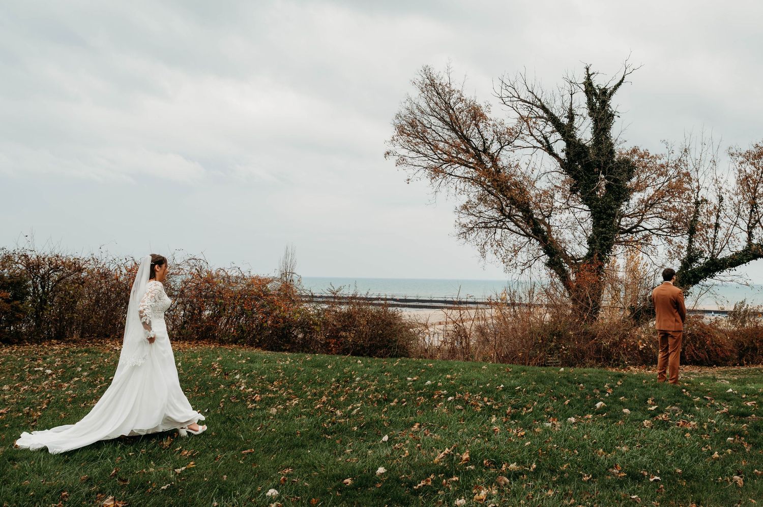 A bride in a flowing white wedding dress walks through an autumn field with bare trees in the background.
