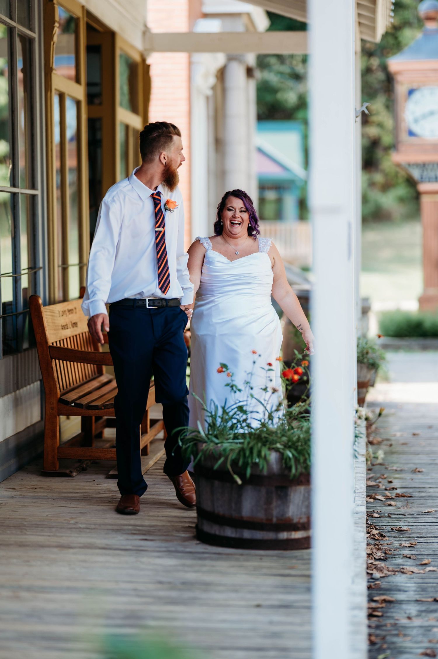 A couple walks together on a covered porch with white columns and wooden planks, with decorative barrel planters nearby.