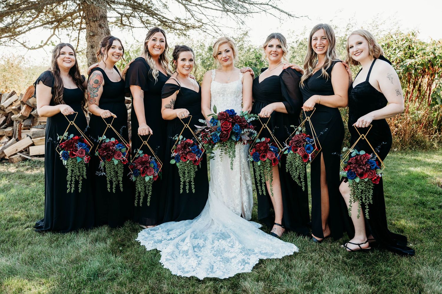 Wedding party poses outdoors with bridesmaids in black dresses holding burgundy bouquets next to bride in white gown.