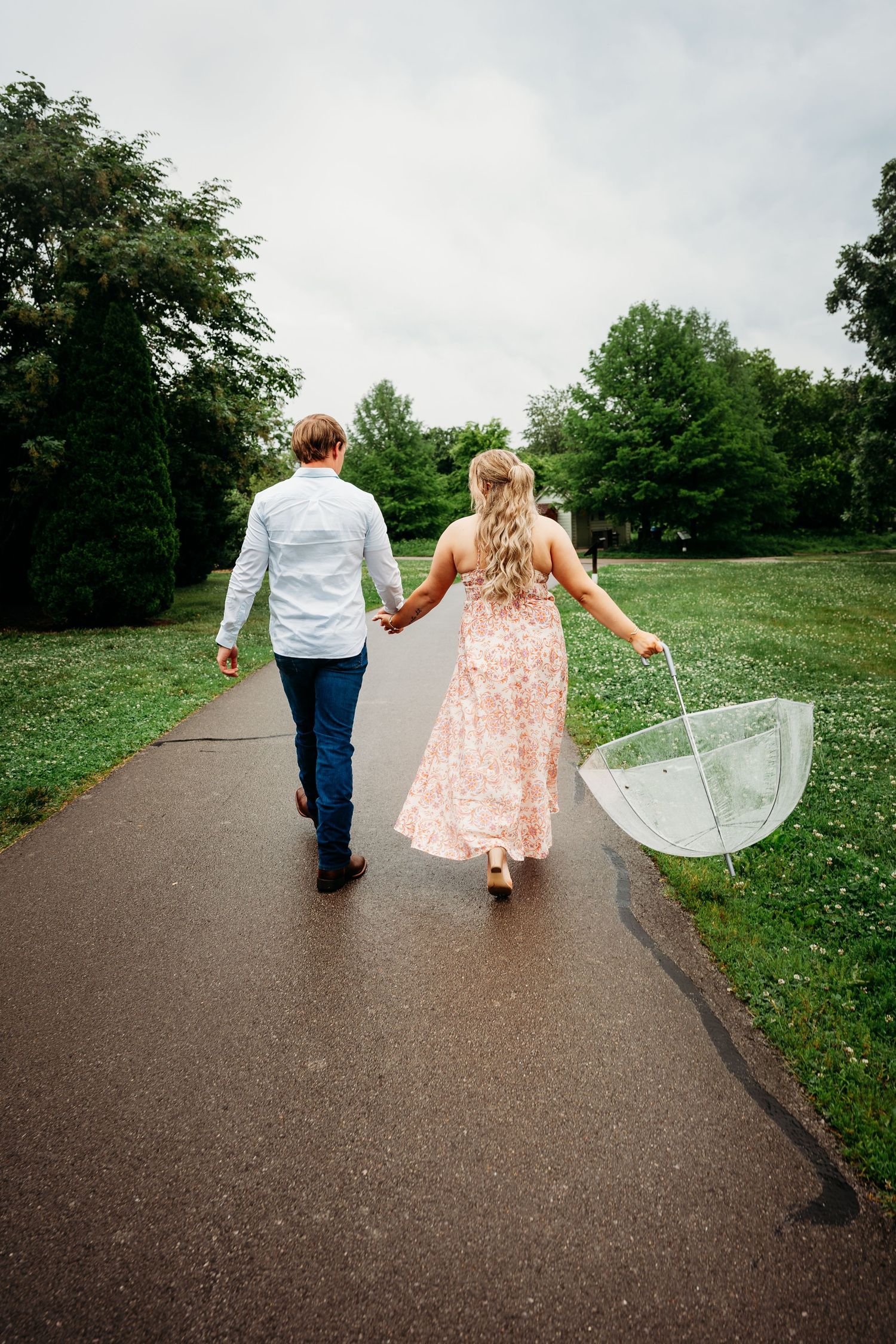 Couple walking hand in hand on a rainy path, woman holding a clear umbrella in a floral pink dress.