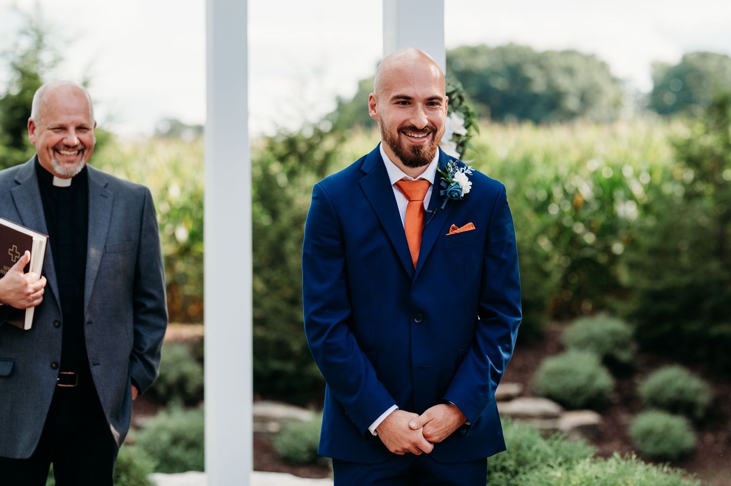 Groom in navy blue suit with orange tie smiling at altar during outdoor wedding ceremony with officiant holding bible.