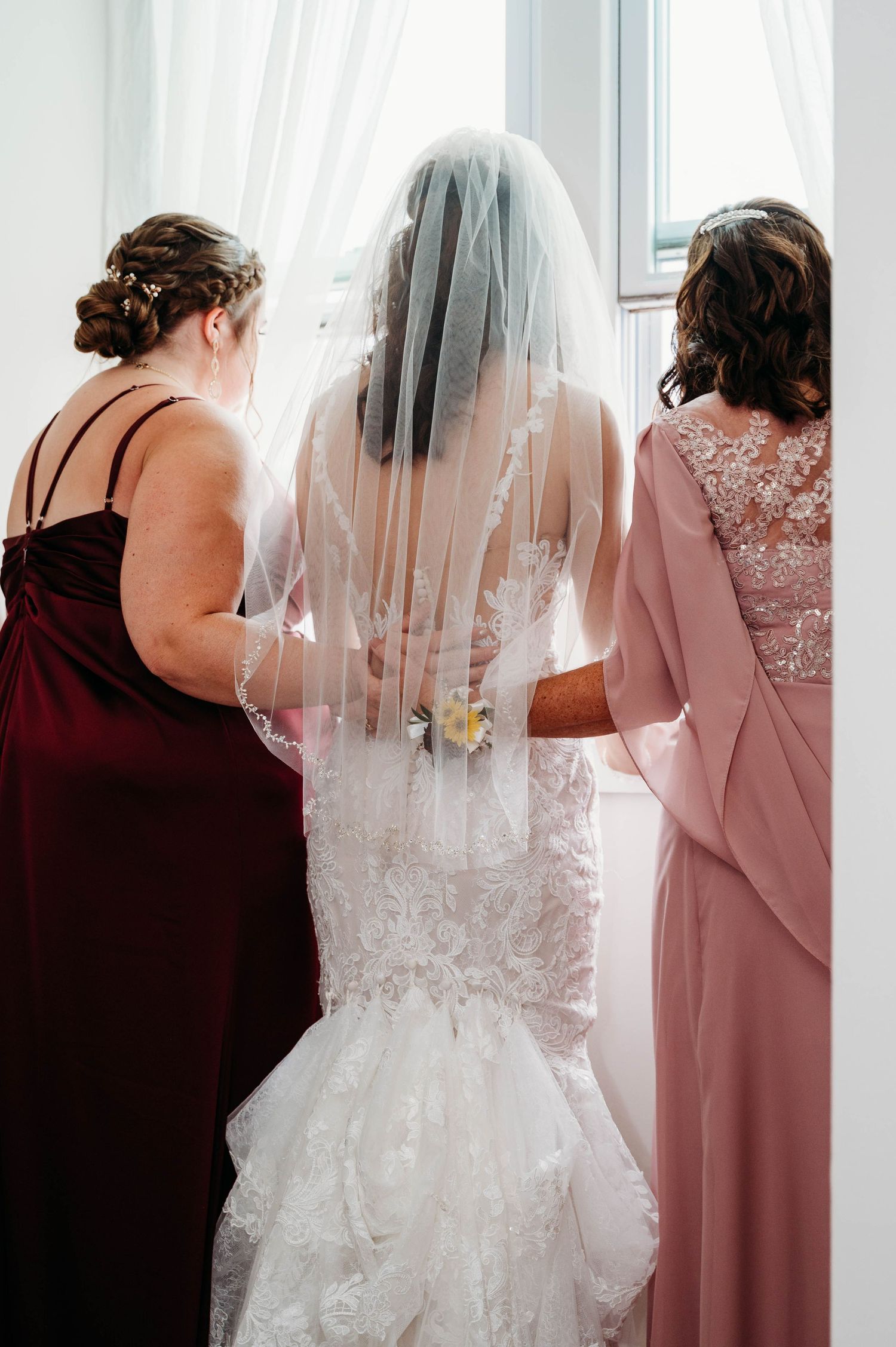 Bride in lace wedding gown and veil flanked by two bridesmaids in burgundy and blush dresses, viewed from behind.
