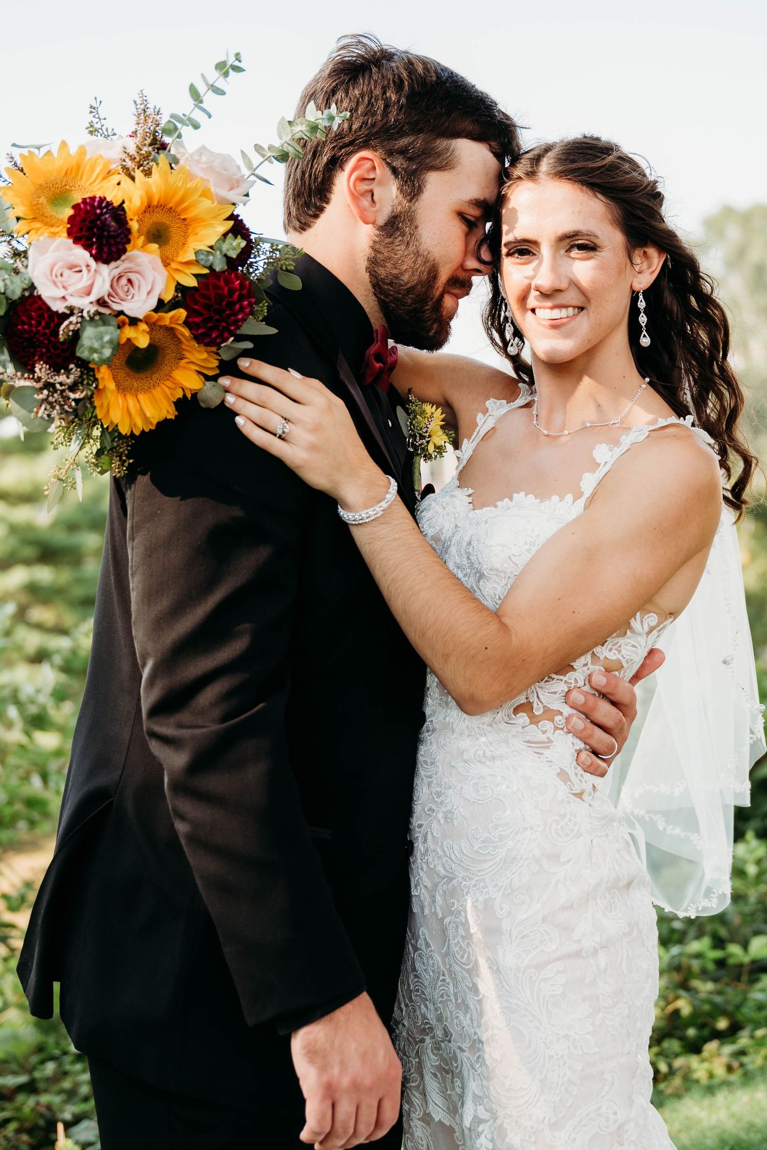 Bride in lace gown and groom in black suit share romantic moment holding sunflower bouquet outdoors.