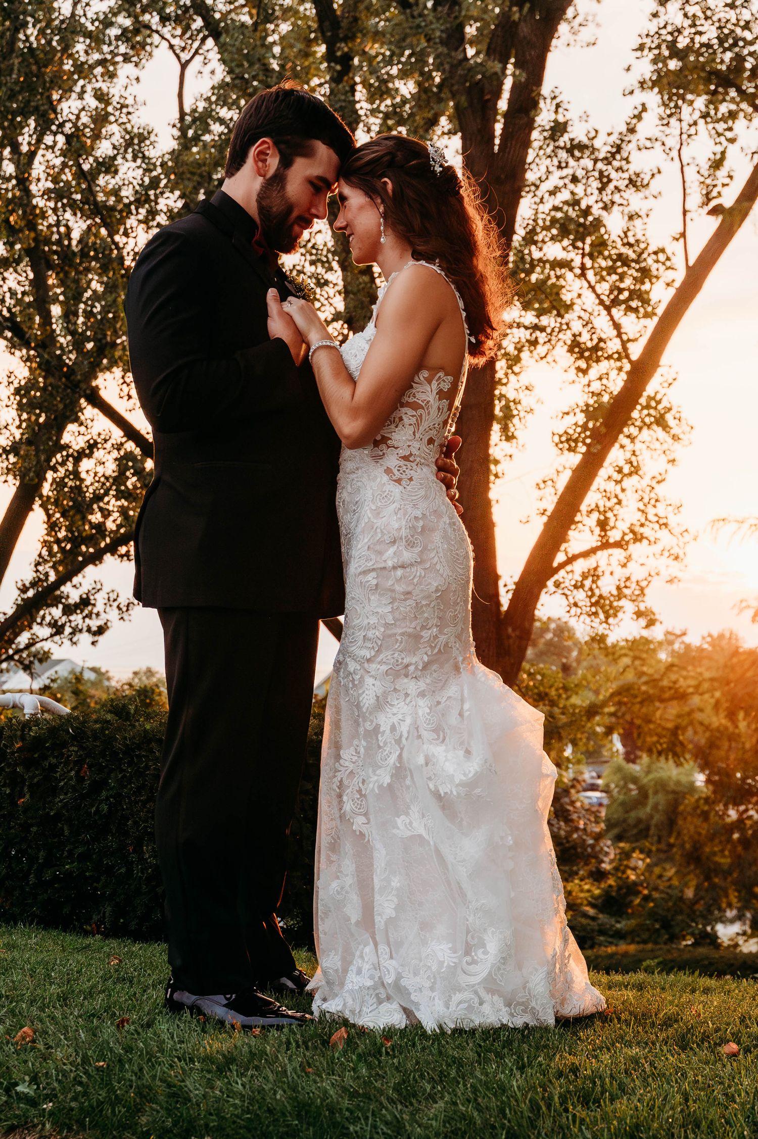 Bride and groom share a romantic sunset moment, foreheads touching, as she wears a lace mermaid gown outdoors.
