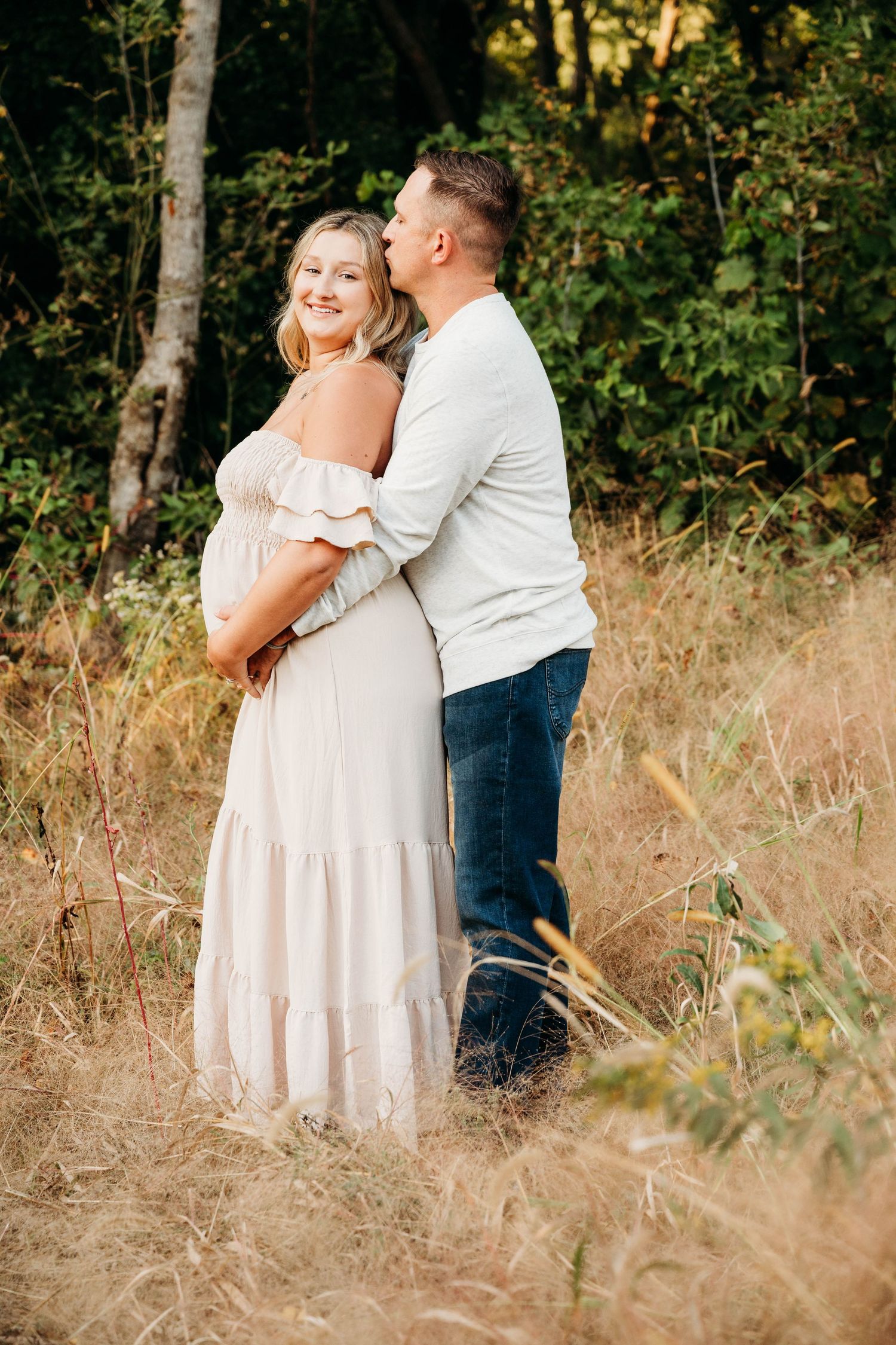 A man embraces a pregnant woman in a white dress outdoors in a golden field with green trees behind them.