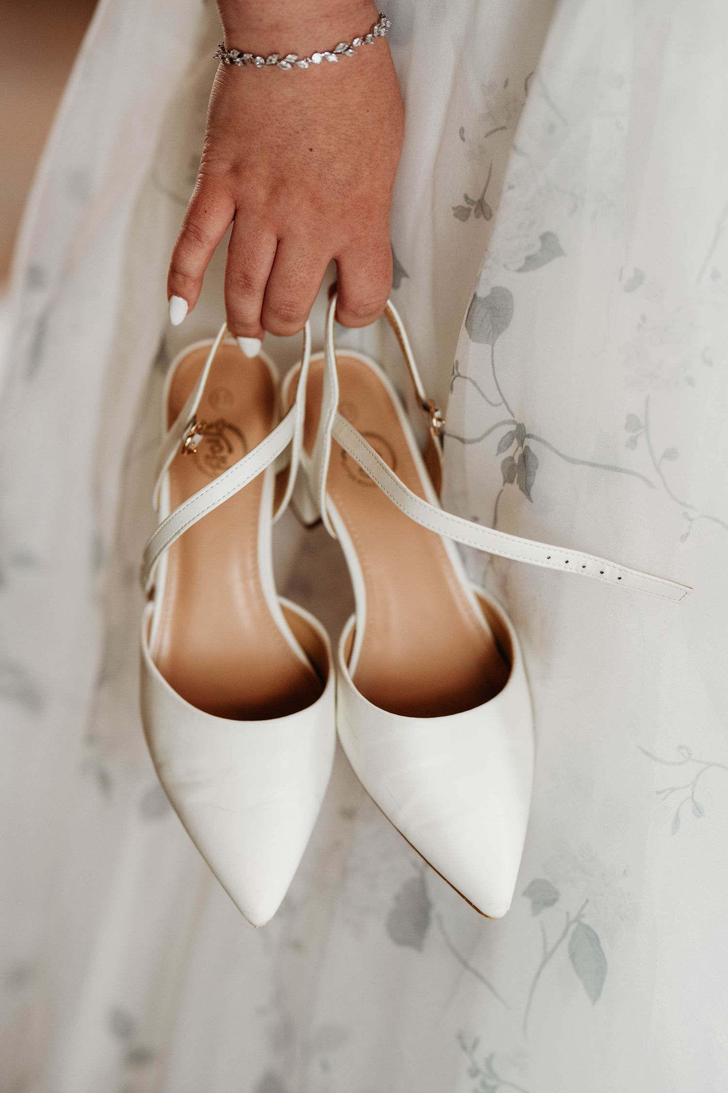 Elegant white pointed-toe bridal flats with delicate ankle straps held against a light floral curtain background.