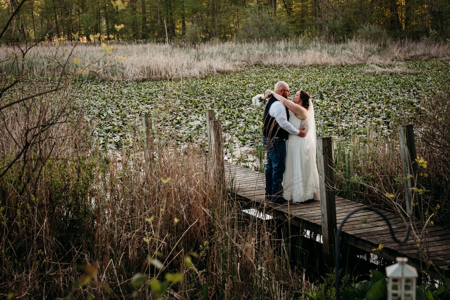 Couple embraces in front of rustic wooden bench surrounded by autumn foliage and tall grass in natural setting.