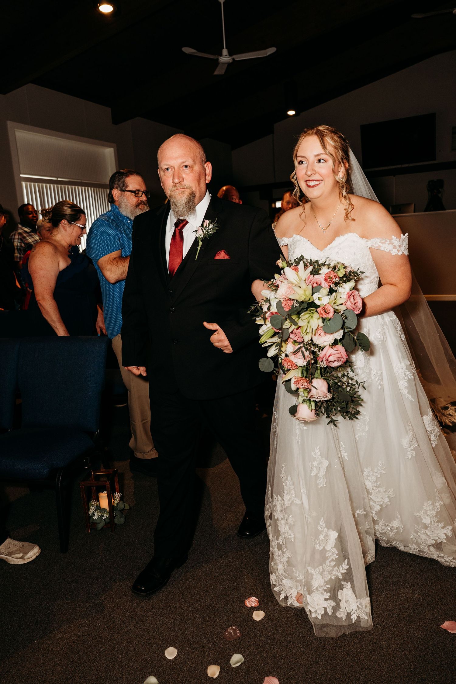 A wedding ceremony scene with a bride in a lace dress holding a pink and white bouquet, walking down the aisle.