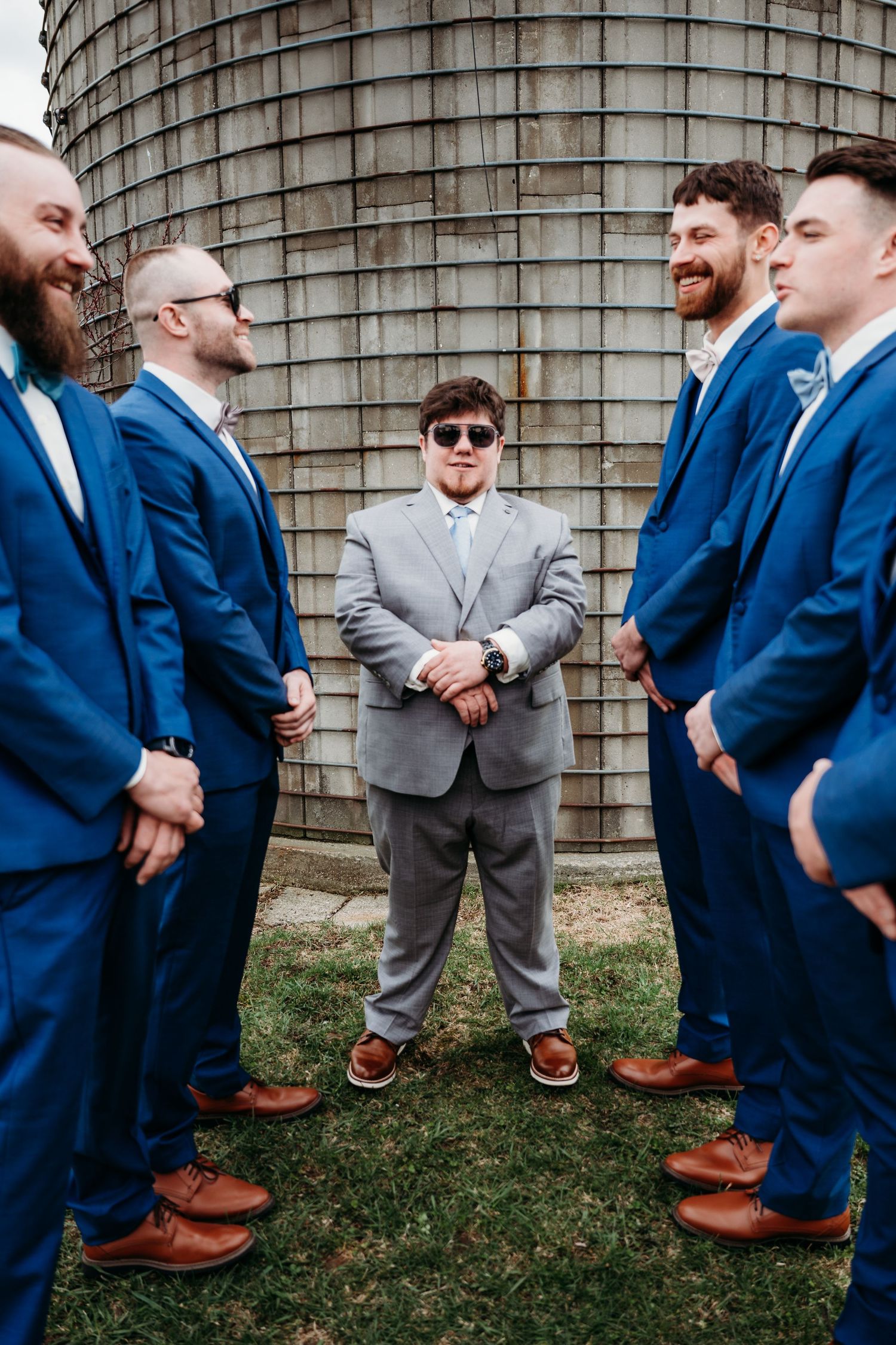 Groomsmen in navy blue suits stand together by a rustic silo with one member wearing a contrasting gray suit.