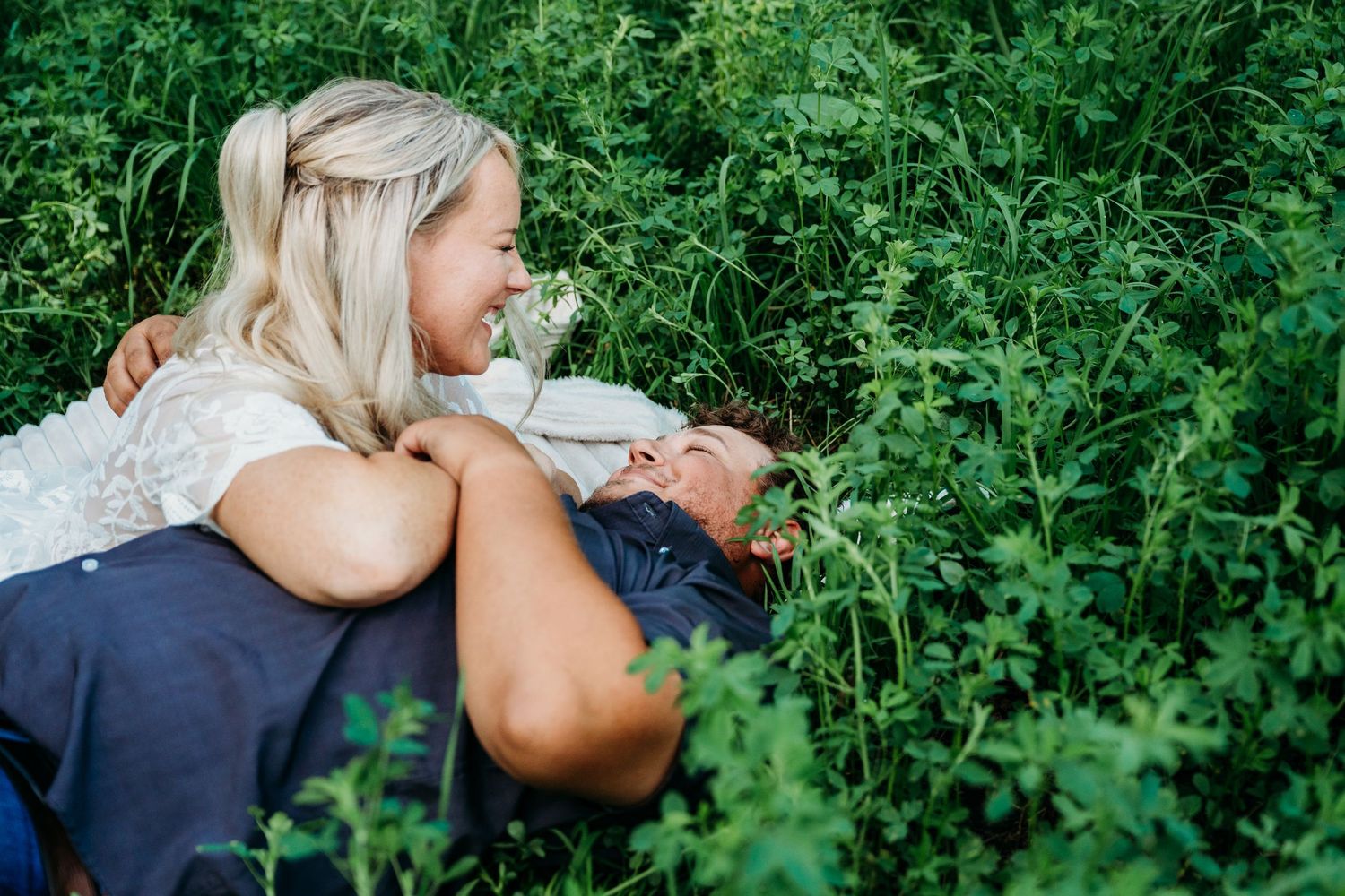 A close-up engagement photo session in a lush green field with tall grasses and wildflowers.