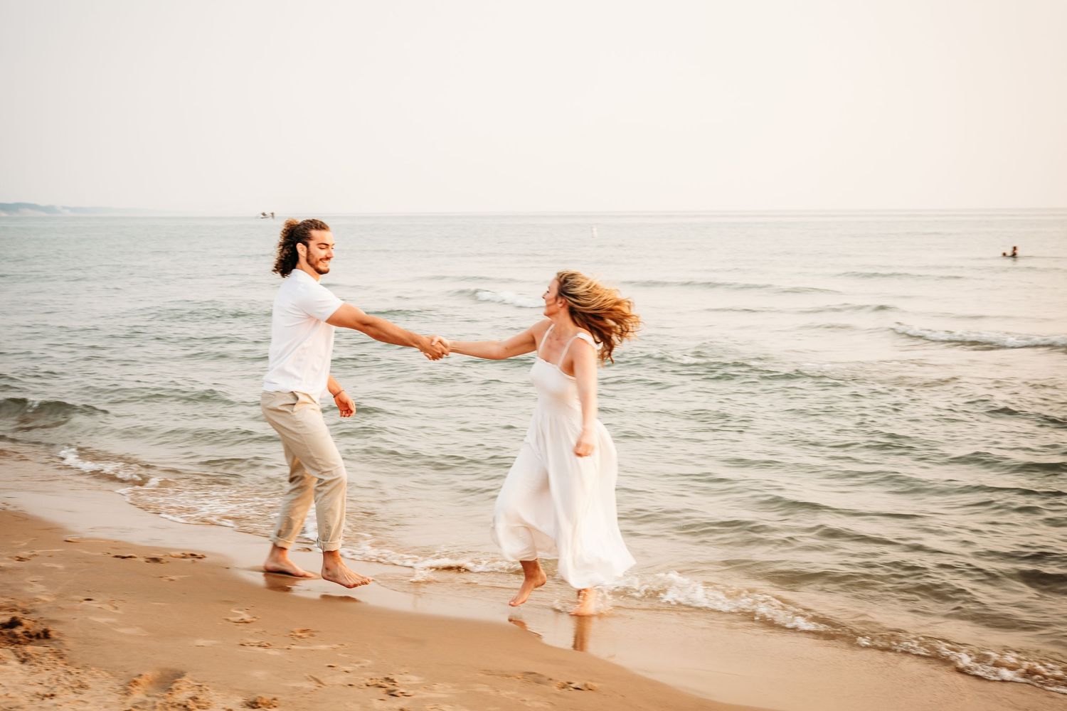 Couple in white clothing holds hands while dancing together on a beach with gentle waves in the background.