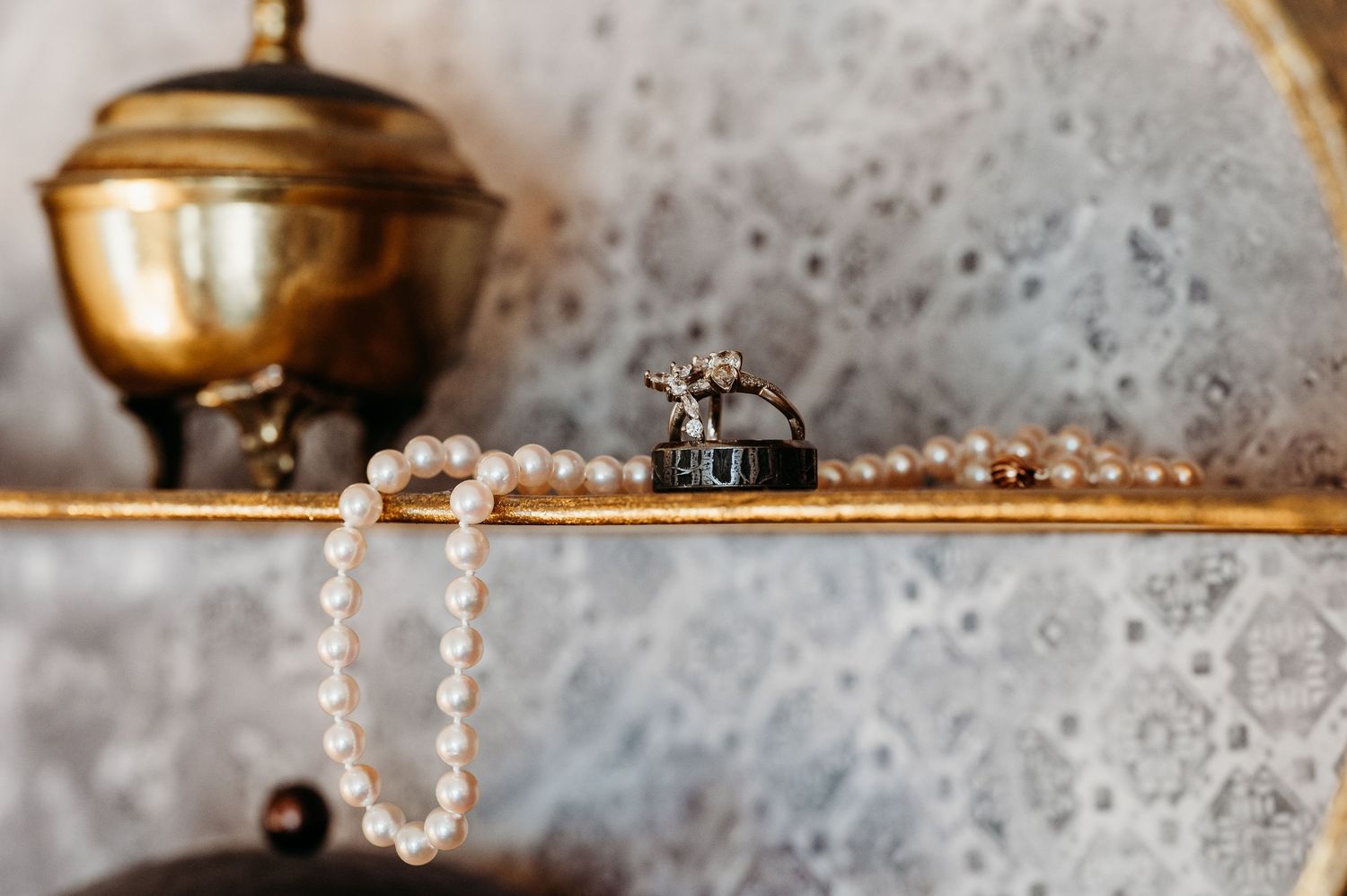 Wedding ring set next to a string of pearls on a vintage gold shelf with an antique brass kettle in soft focus.
