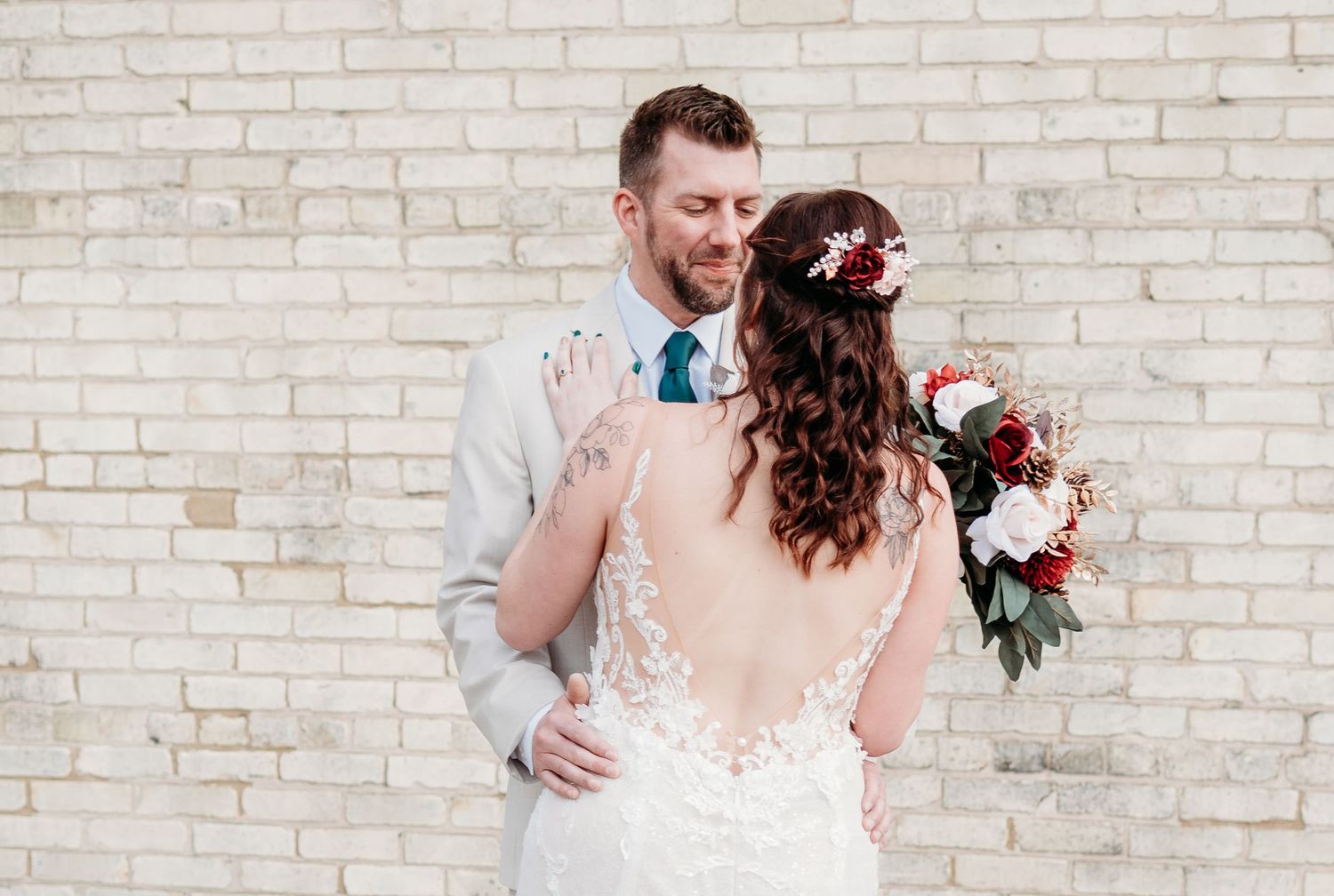 A couple in wedding attire embrace against a white brick wall, with the bride holding a red and white bouquet.