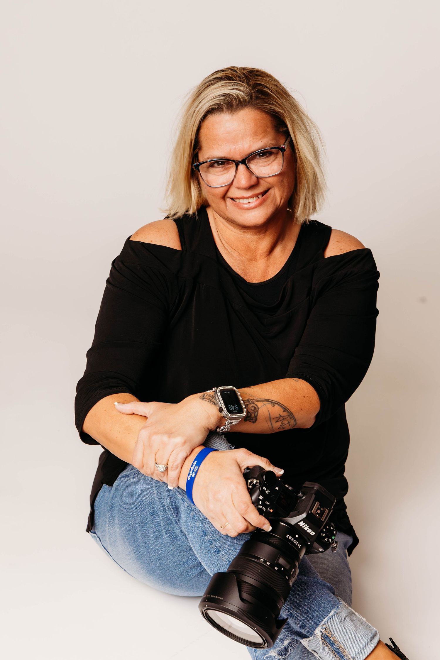 Professional photographer in black top and jeans poses with camera in studio against white backdrop.