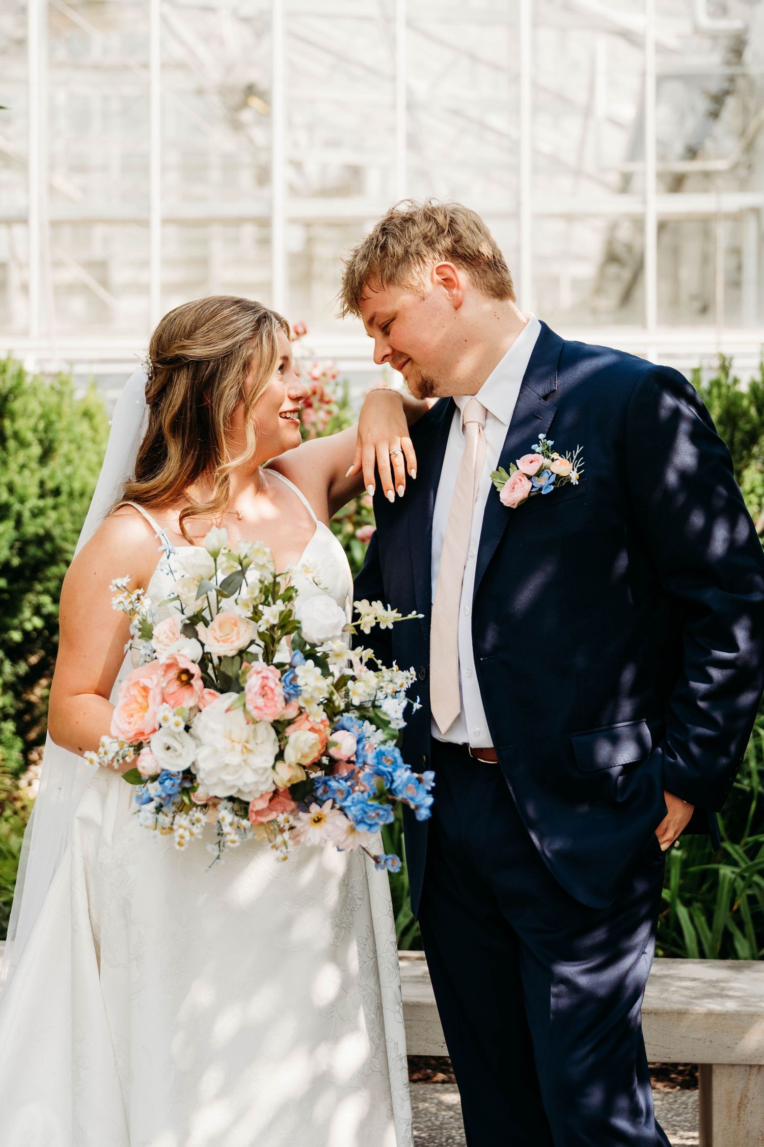 Bride and groom share a romantic moment outdoors, she holds a pastel bouquet, he wears a navy suit with floral boutonniere.
