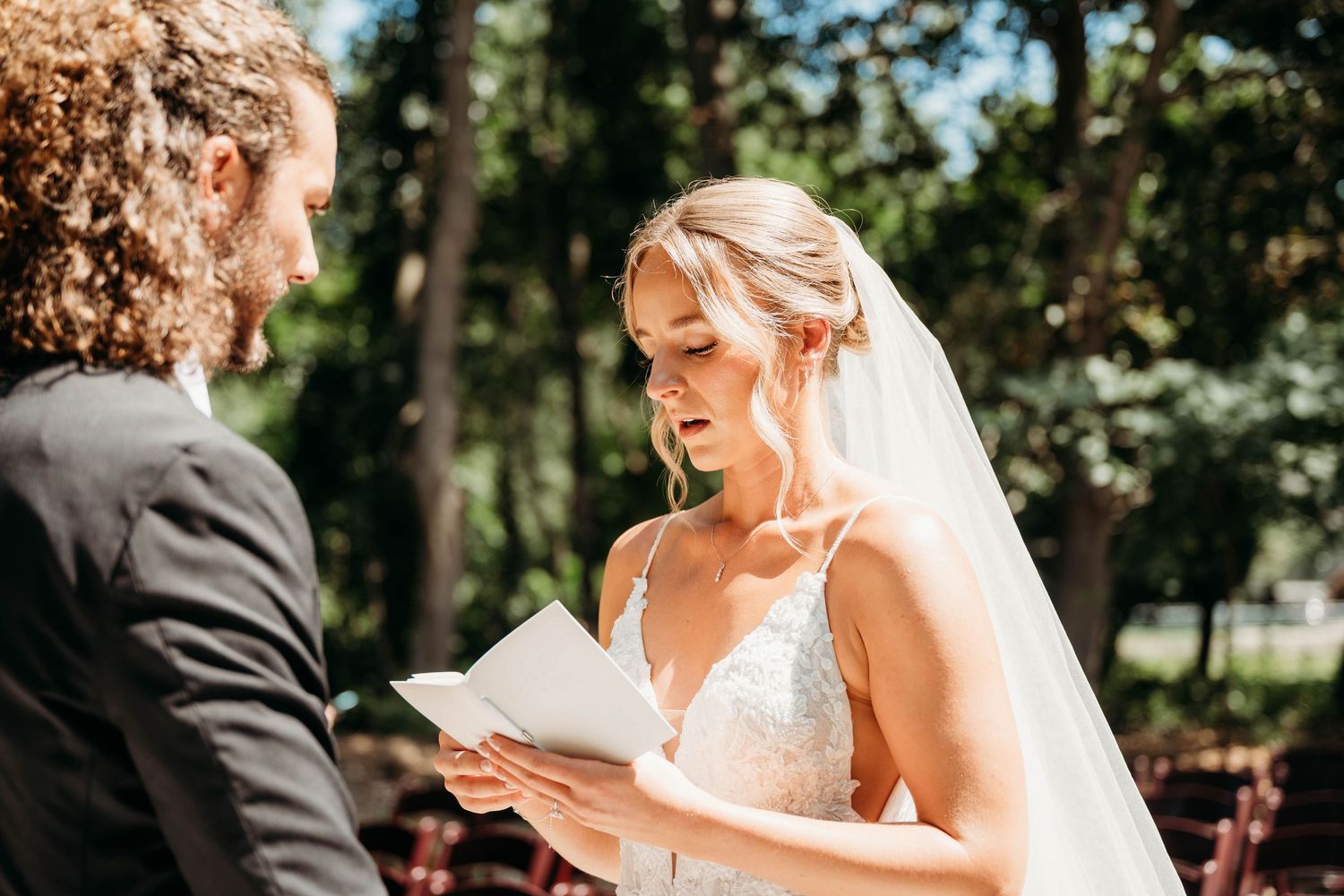 Bride in lace gown and veil reads wedding vows outdoors during romantic garden ceremony.