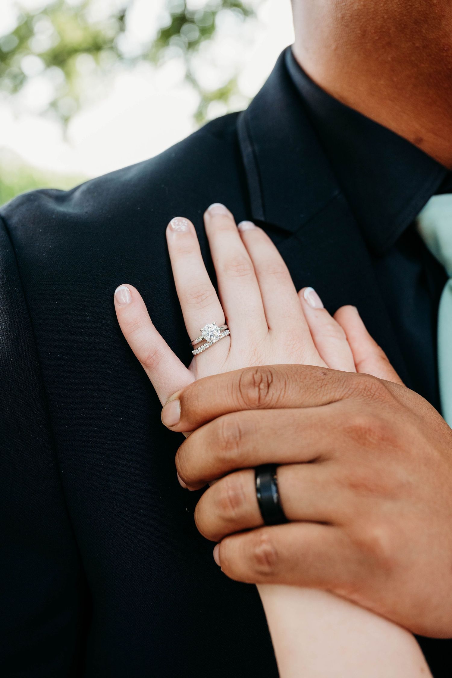 Close-up of bride and groom hands showing diamond engagement ring and black wedding band on dark suit.
