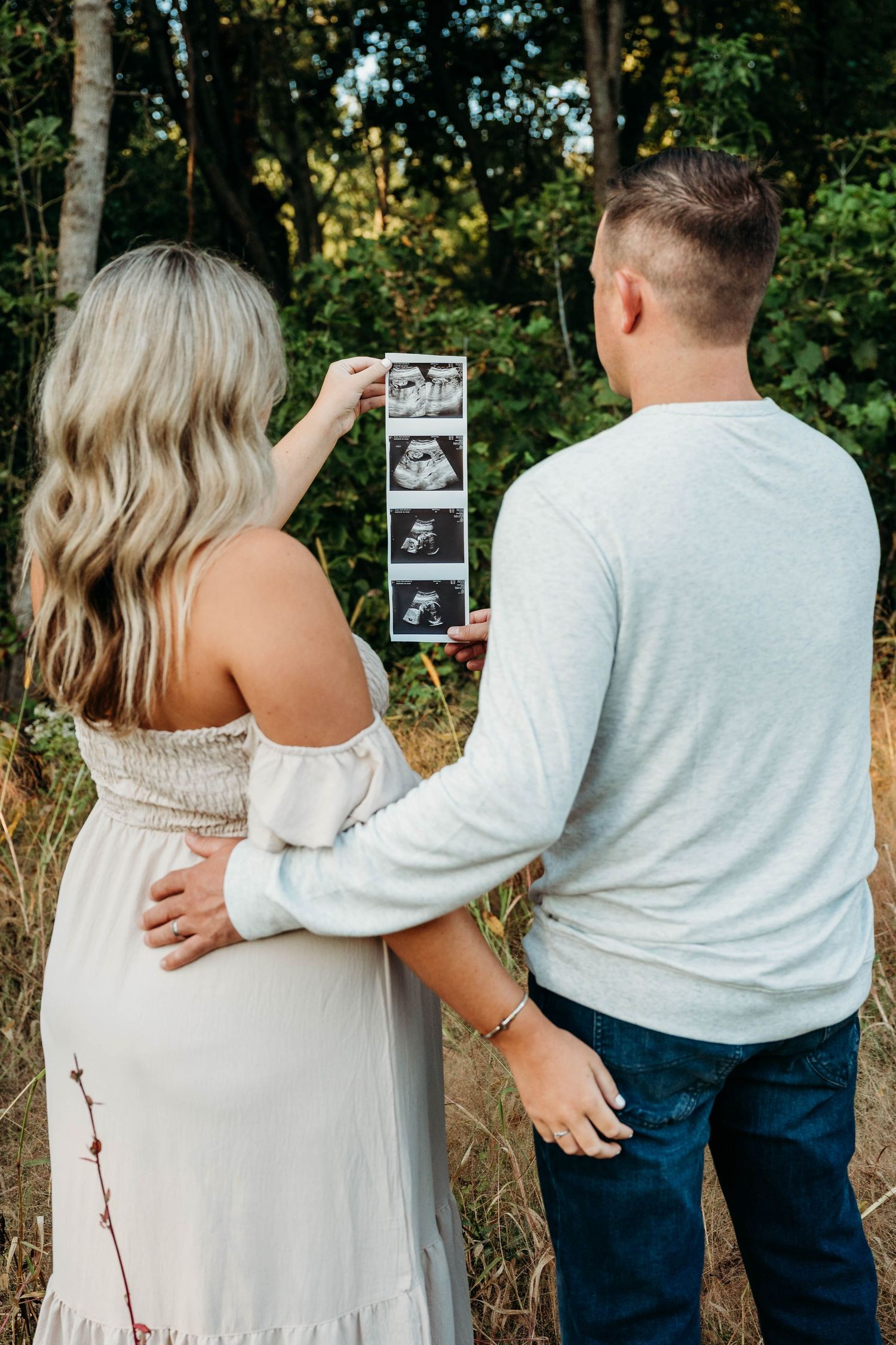 A couple in casual attire holds up ultrasound photos during a pregnancy announcement photoshoot outdoors.